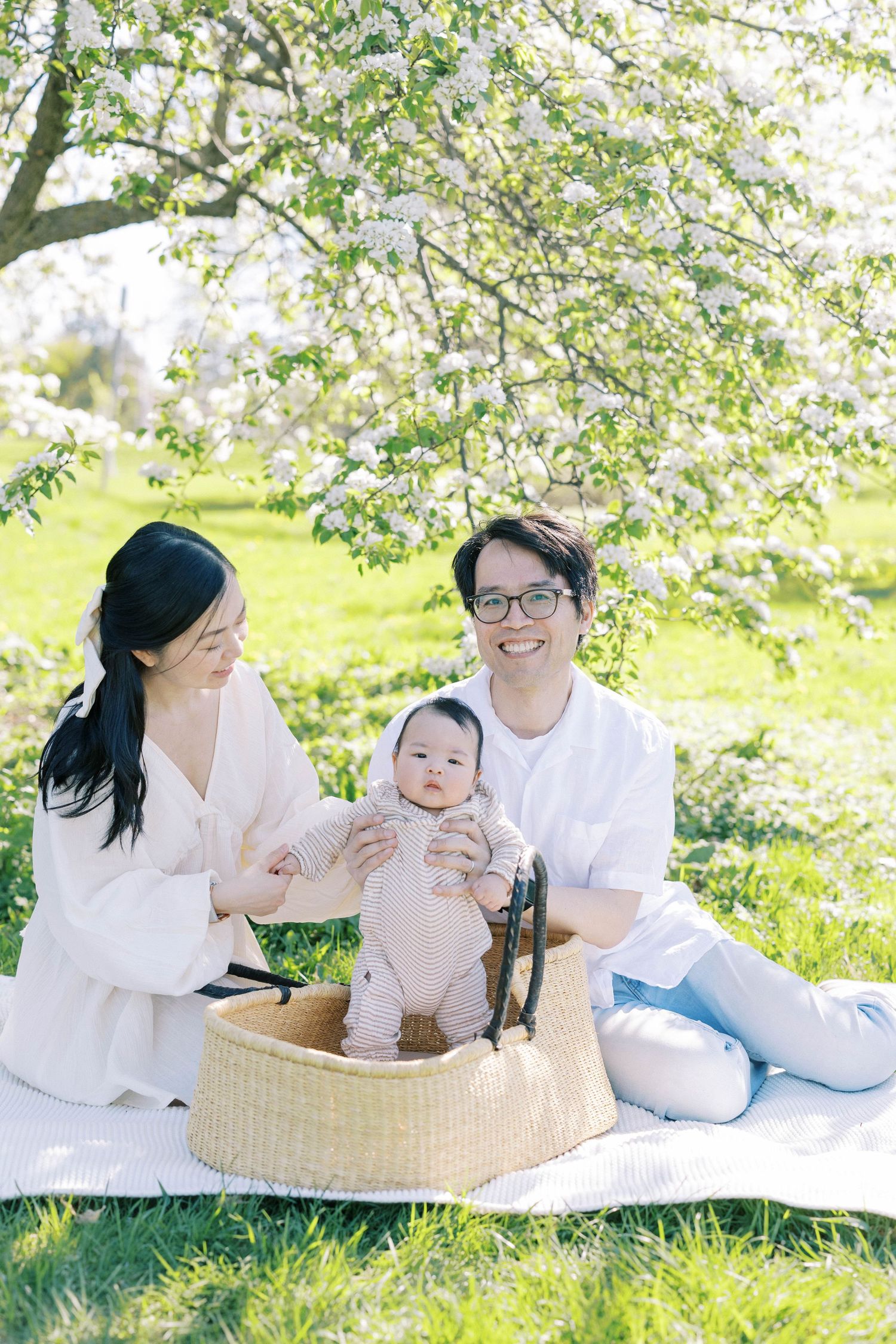 Happy Asian family with mother, father, and baby in Moses basket sitting on blanket under flowering spring tree.