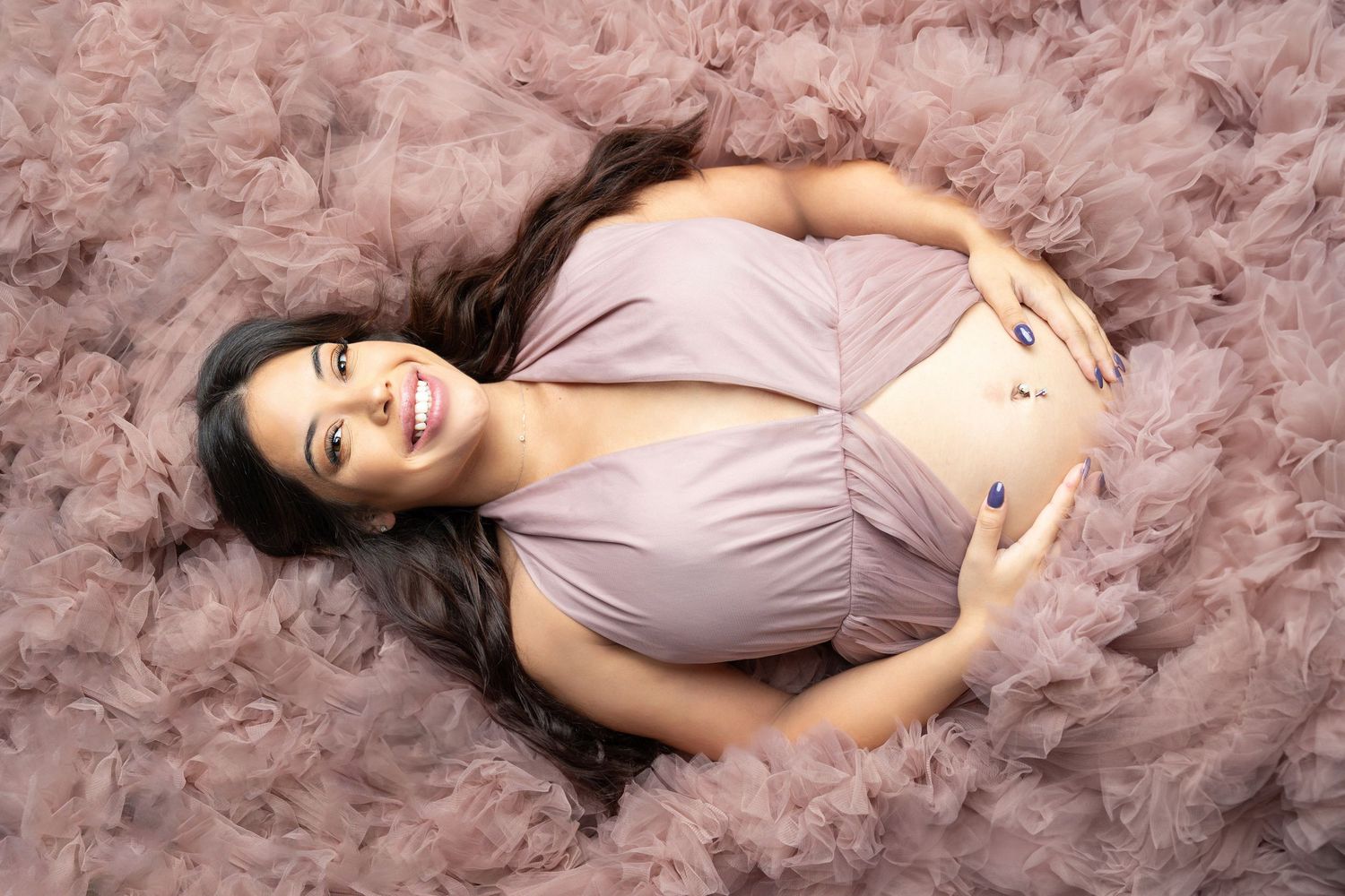 Joyful pregnant woman lying on pink tulle fabric, smiling while holding her baby bump.