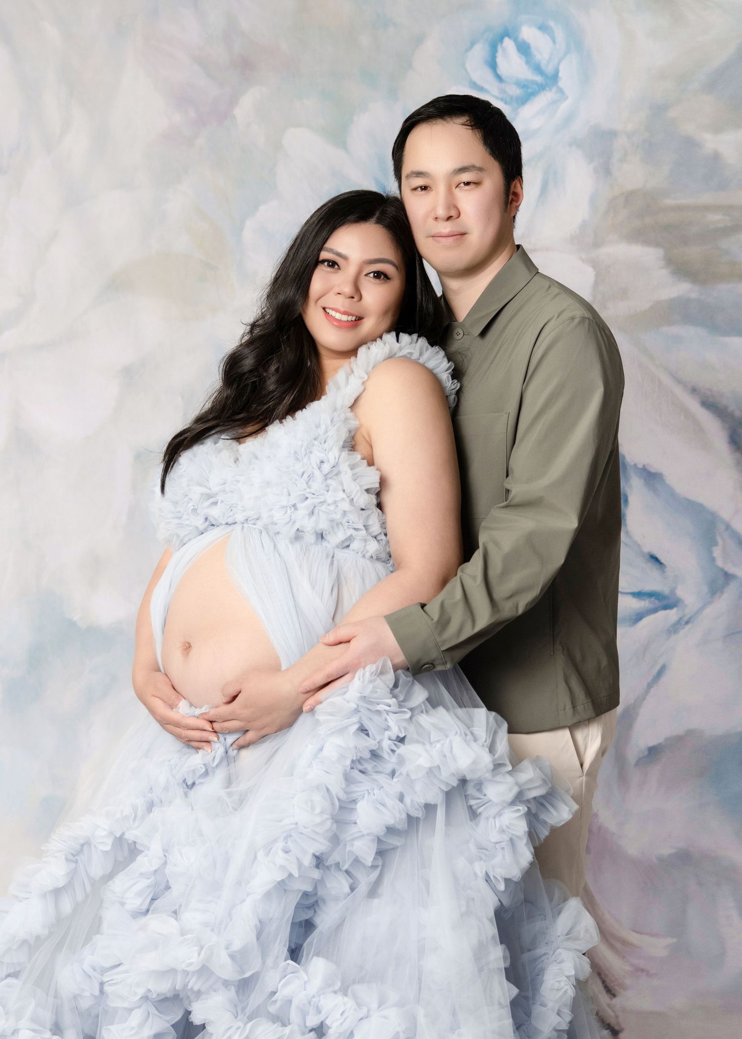 Pregnant woman in ruffled blue gown poses with partner in olive shirt for maternity photo shoot.