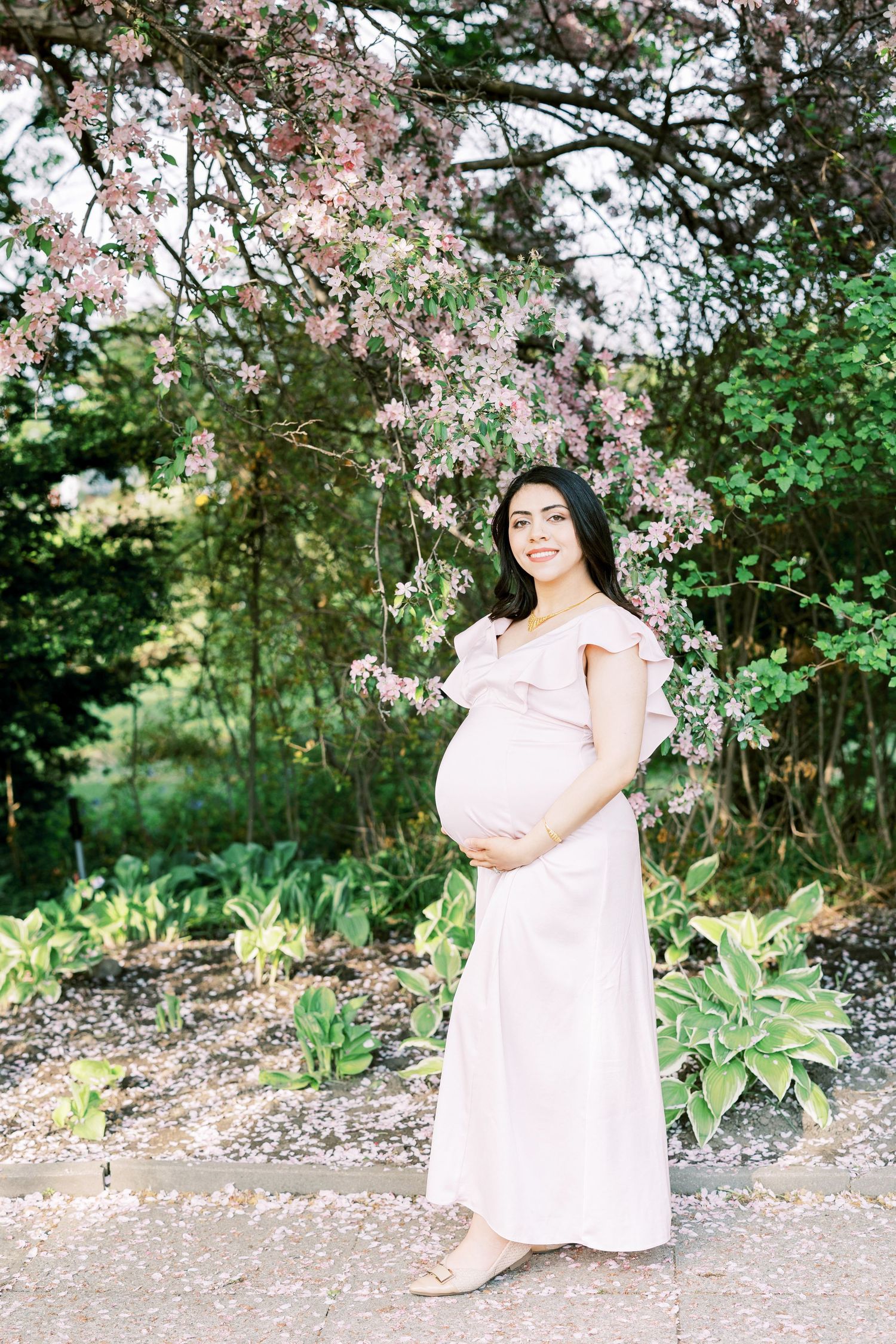 Radiant pregnant woman in blush pink off-shoulder gown cradling baby bump under pink flowering tree in garden.