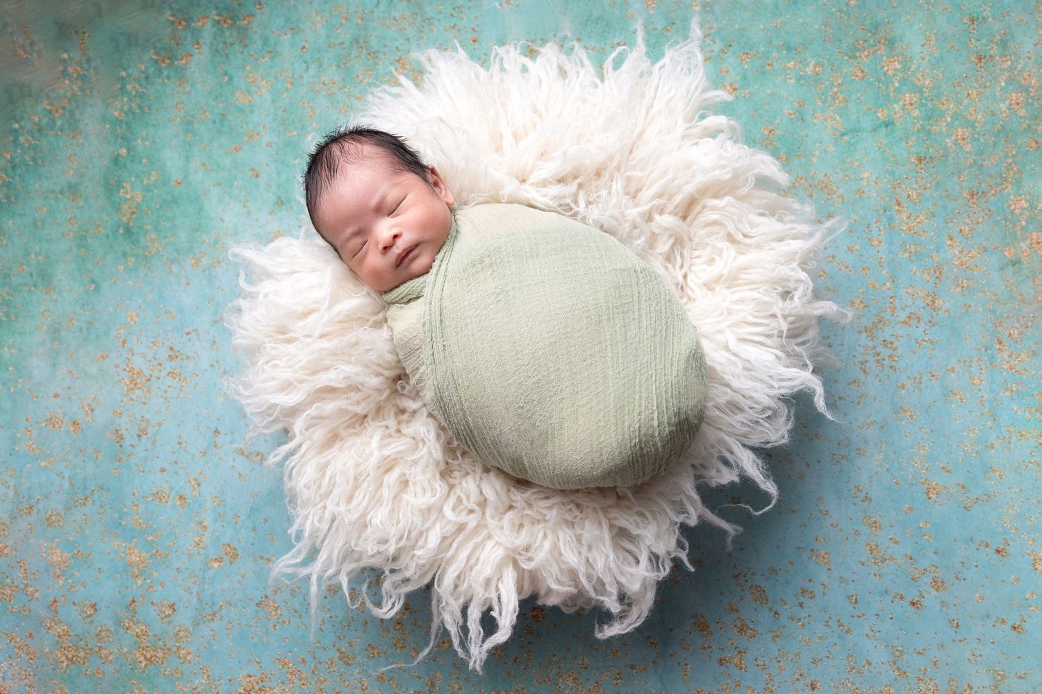 Sleeping newborn swaddled in green wrap nestled in fluffy white bowl on teal background.