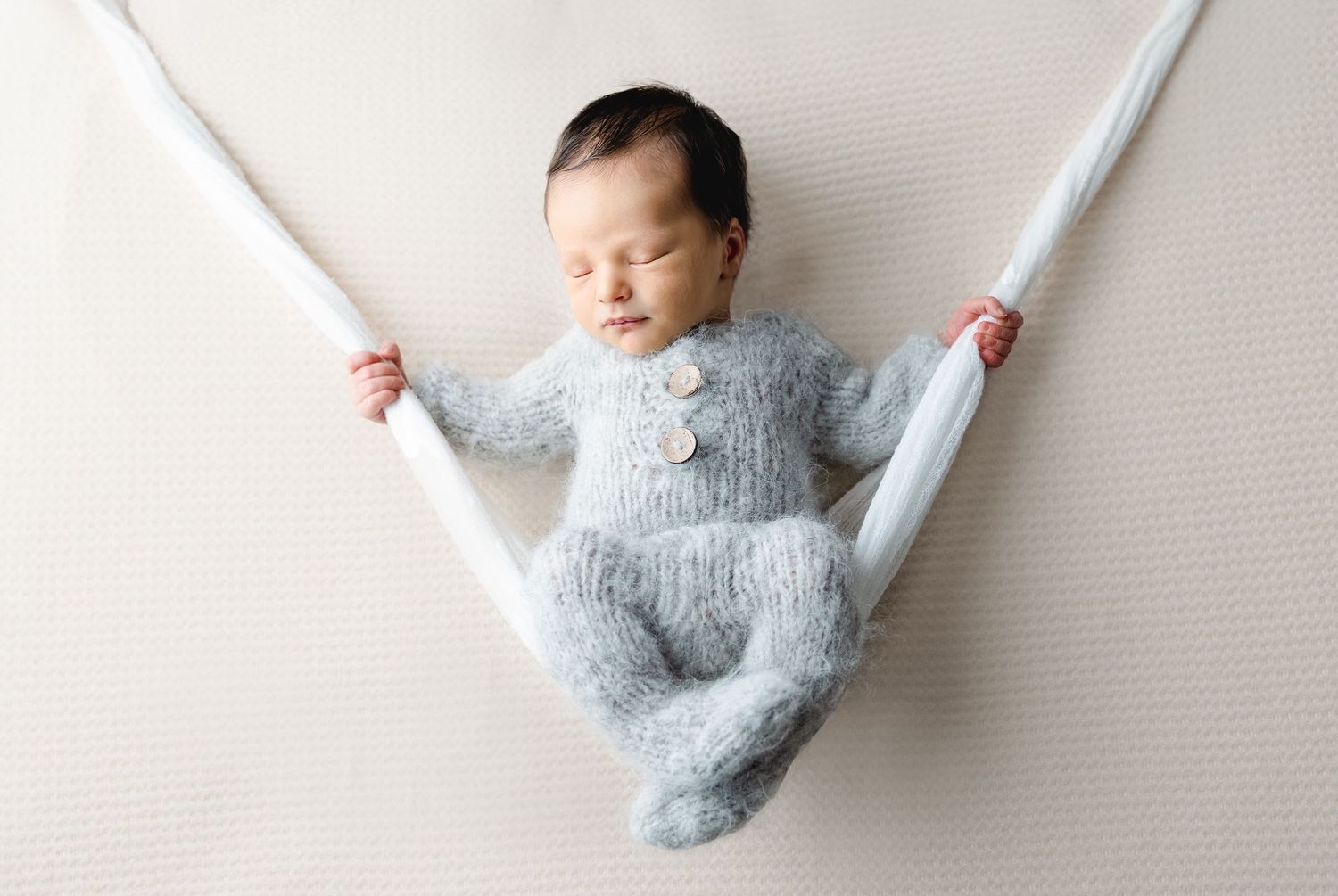 Peaceful newborn baby in grey knit romper sleeping in fabric hammock on white background.