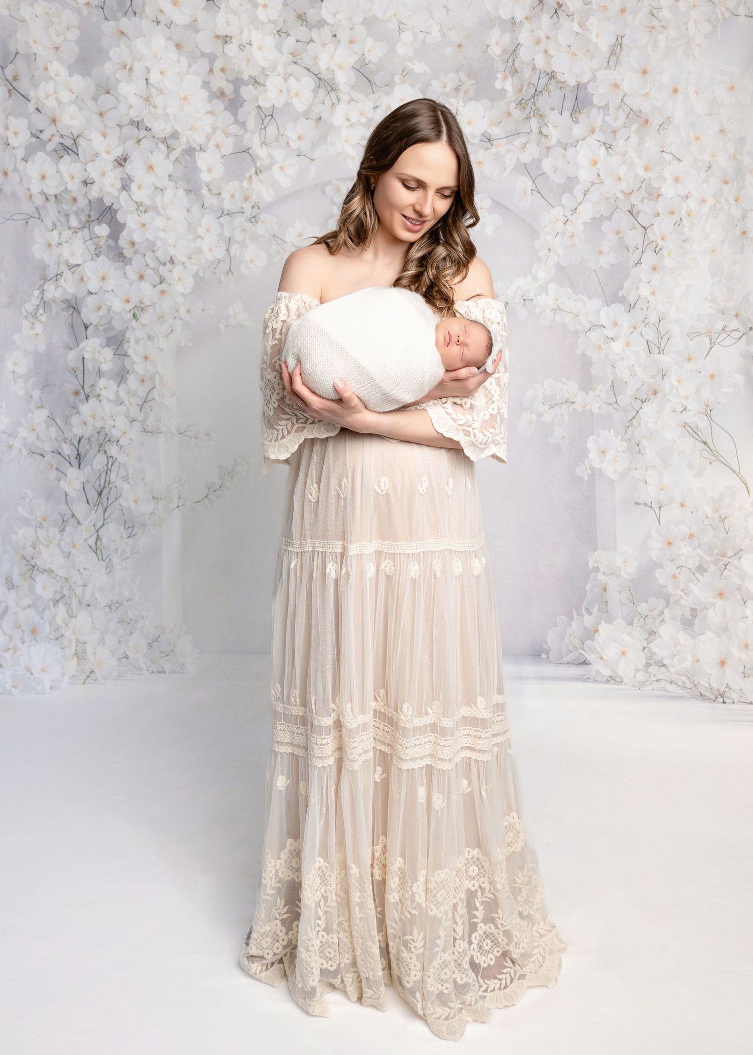 A mother in a white lace gown holds her swaddled newborn baby against a floral backdrop.