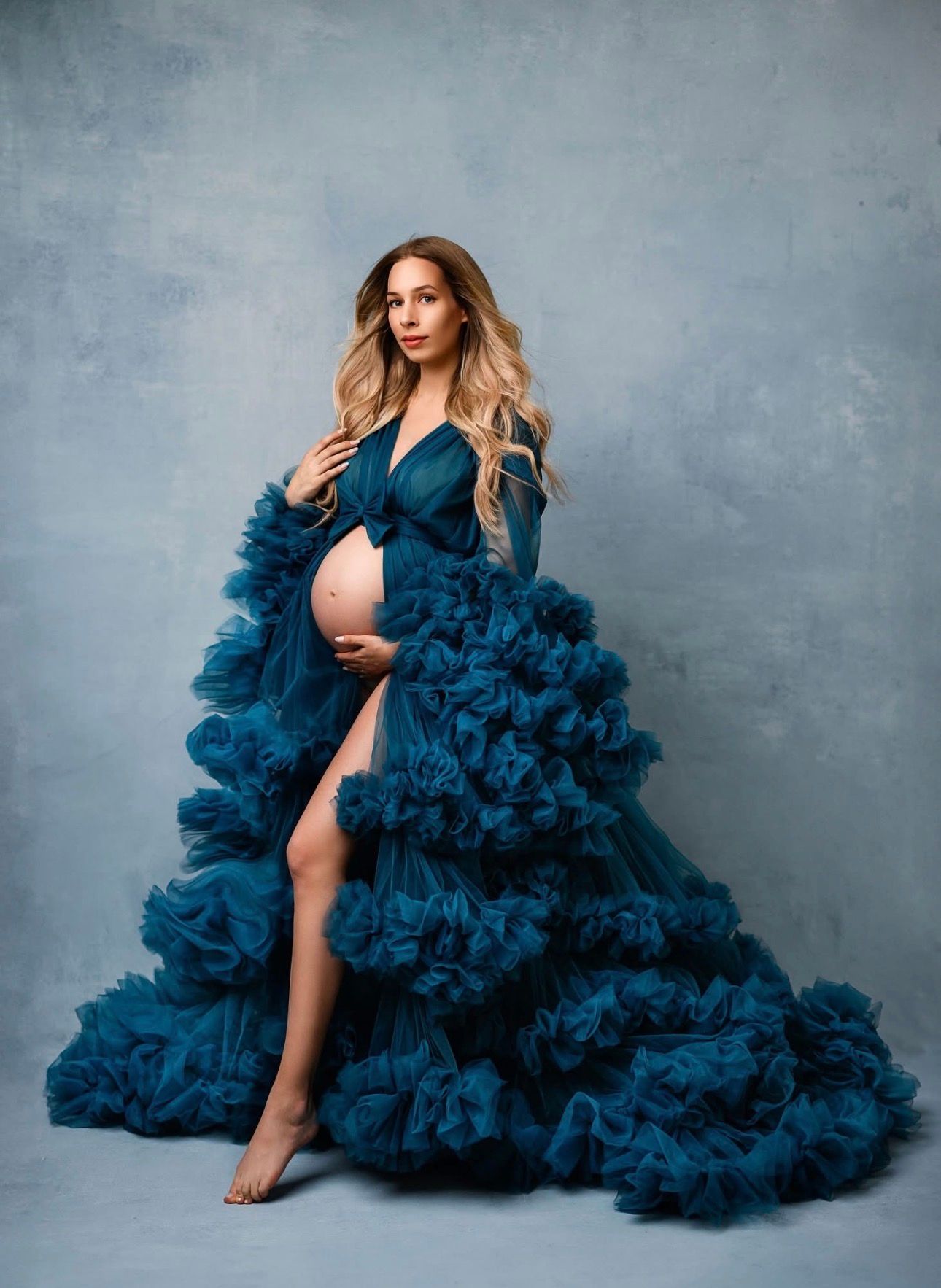 Pregnant woman in elegant teal ruffled tulle maternity gown posing gracefully in a professional studio photo shoot.