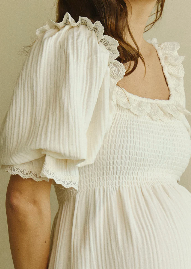 White smocked bodice dress with puff sleeves, lace trim details, and square neckline in textured cotton fabric.