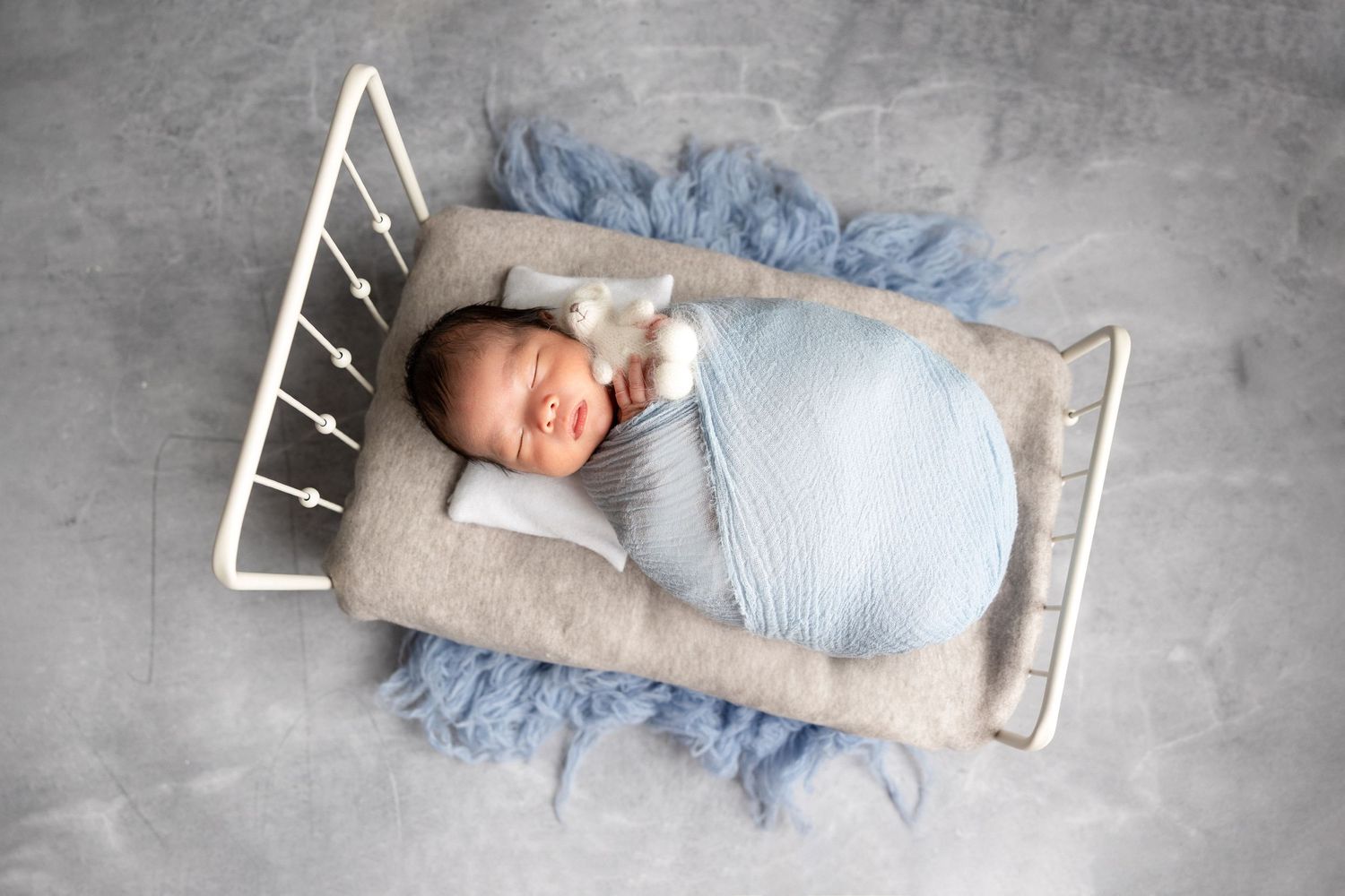 Peaceful newborn baby swaddled in light blue wrap sleeping on tiny white metal bed with blue fur.