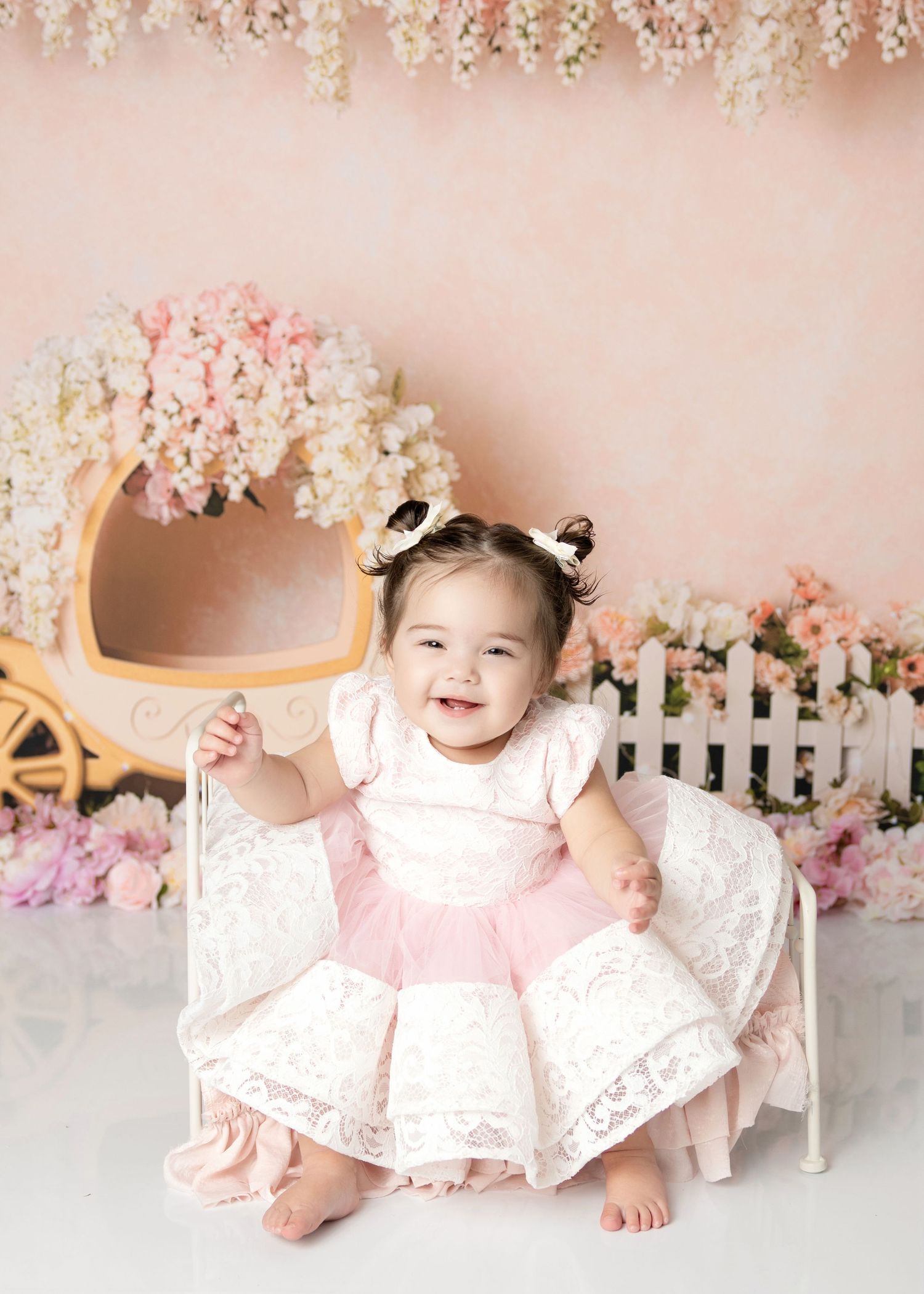 Happy baby girl in pink and white lace dress smiles on white bench with floral carriage backdrop.