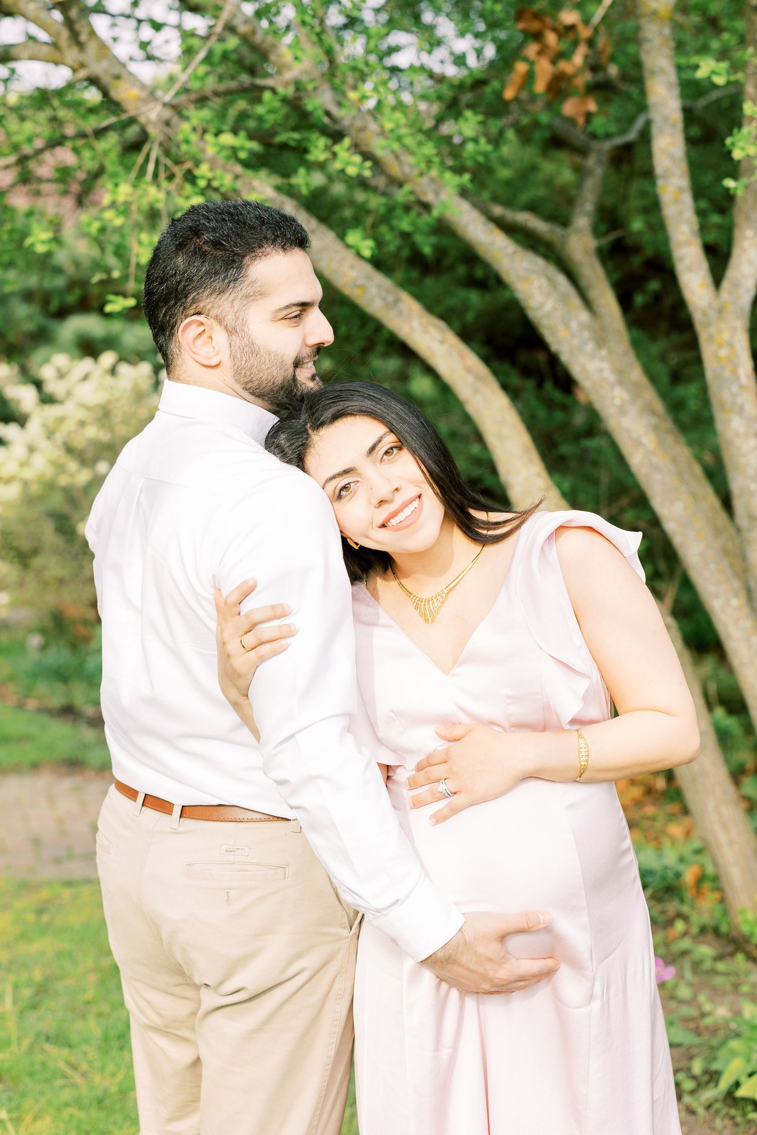 Pregnant woman in pink dress embraces man in white shirt outdoors in lush garden during maternity photo session.