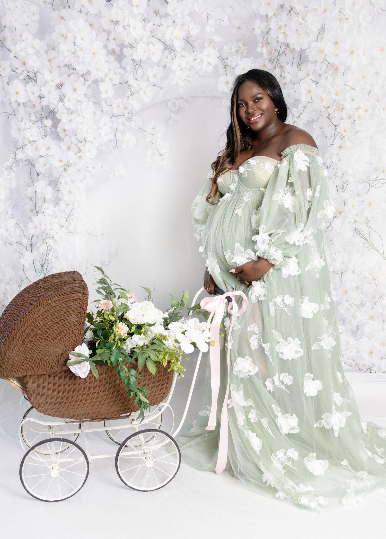 Pregnant woman in sage green tulle gown posing with vintage wicker baby carriage and flowers.