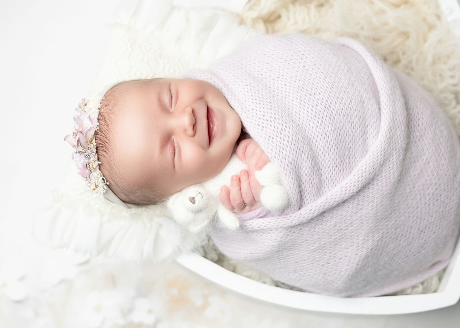 Sleeping newborn baby swaddled in lavender knit wrap with floral headband in a white bowl.