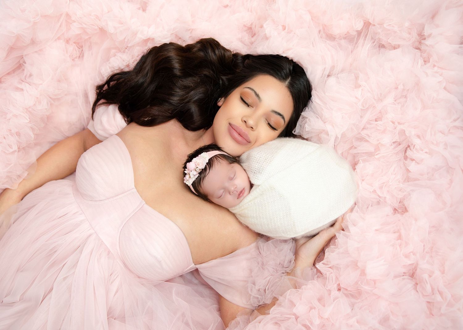 Beautiful mother in pink tulle gown lying with sleeping swaddled newborn on ruffled backdrop.