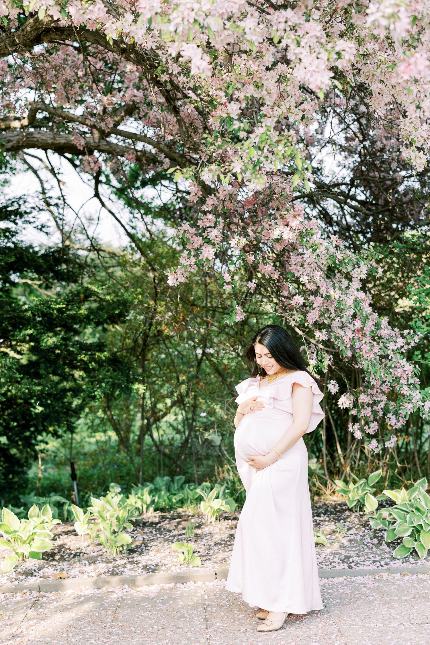 Pregnant woman in pink off-shoulder gown stands under blooming cherry blossom tree in lush garden.