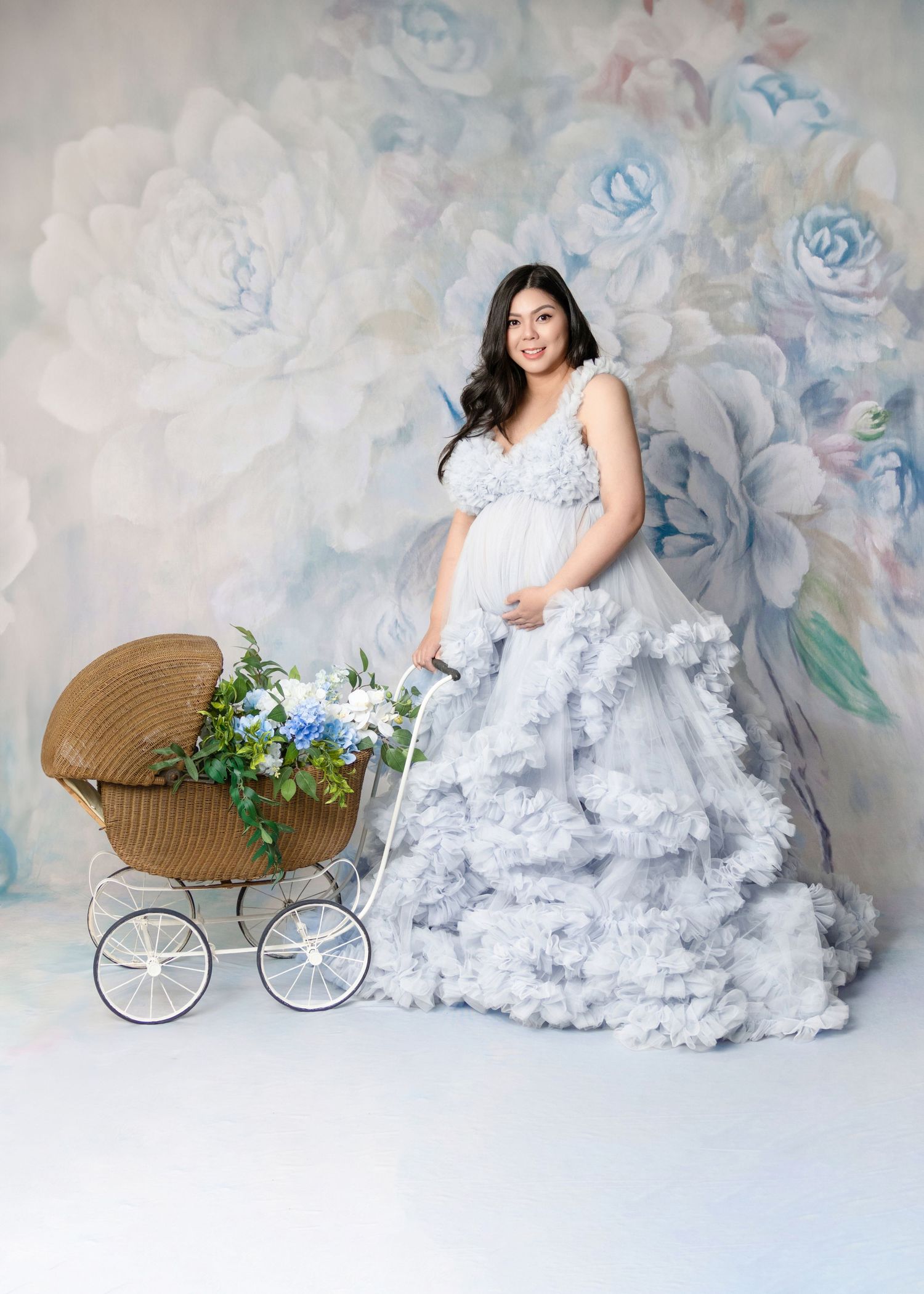 Pregnant woman in white ruffled gown poses with vintage wicker pram and floral backdrop.