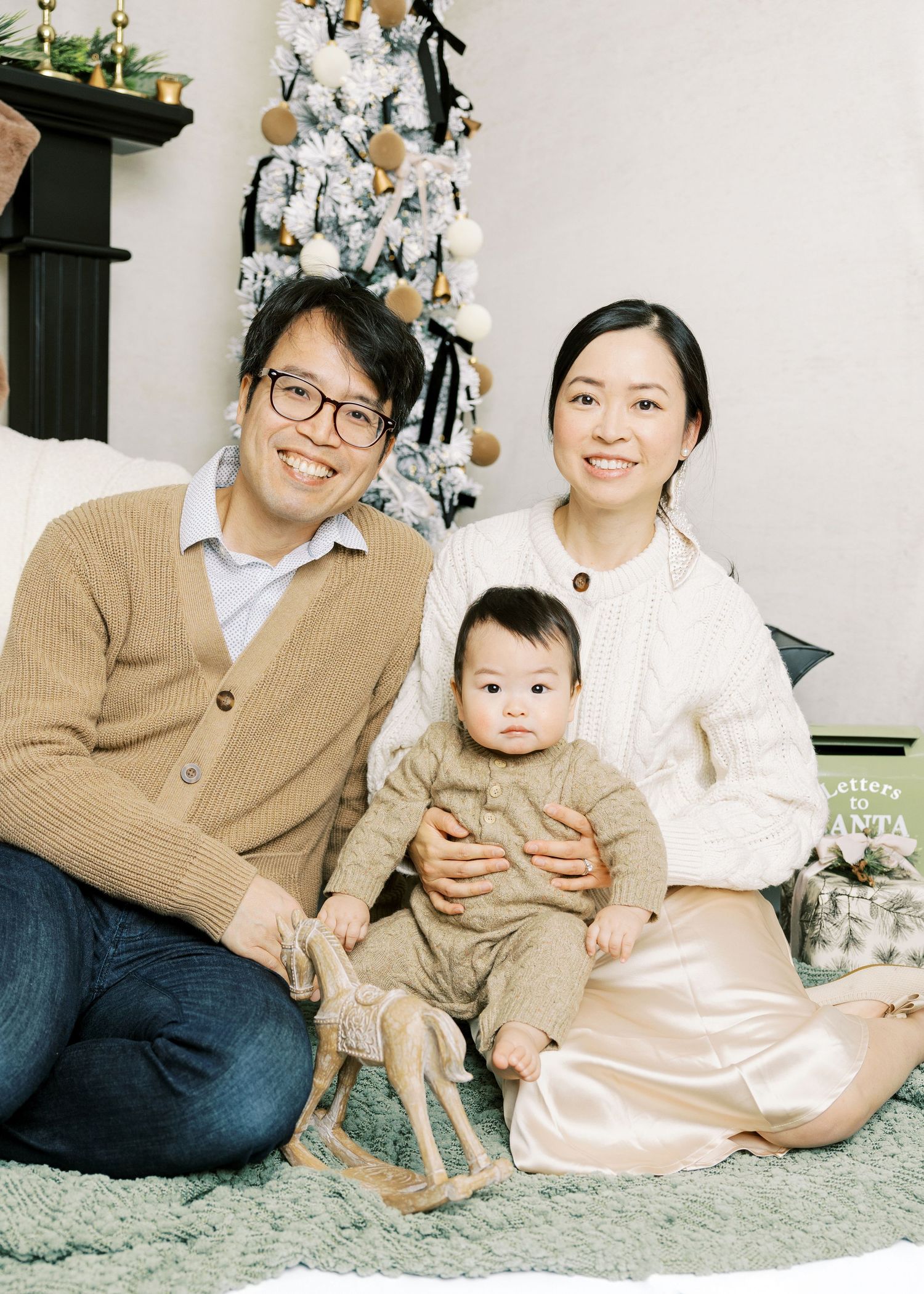 Asian family Christmas photo with parents and baby in front of decorated white tree.