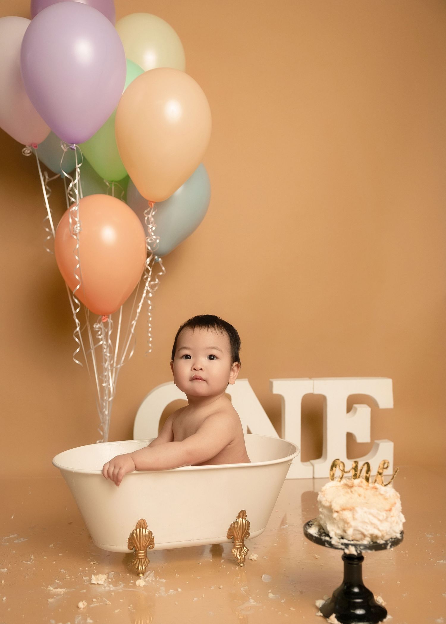 Baby sitting in white clawfoot tub with pastel balloons and birthday cake on beige backdrop.