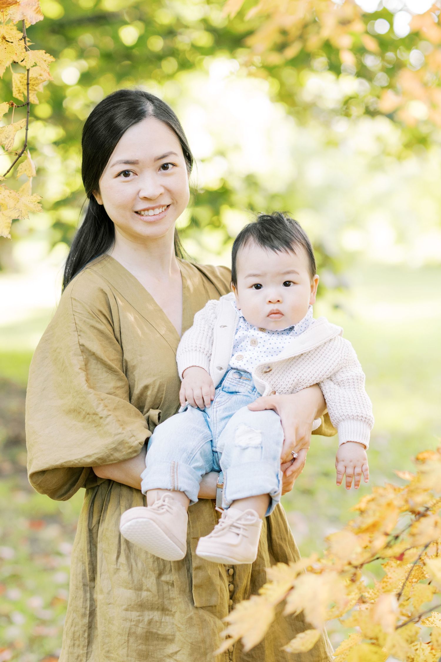 A smiling mother in an olive dress holds her baby girl among autumn leaves outdoors.