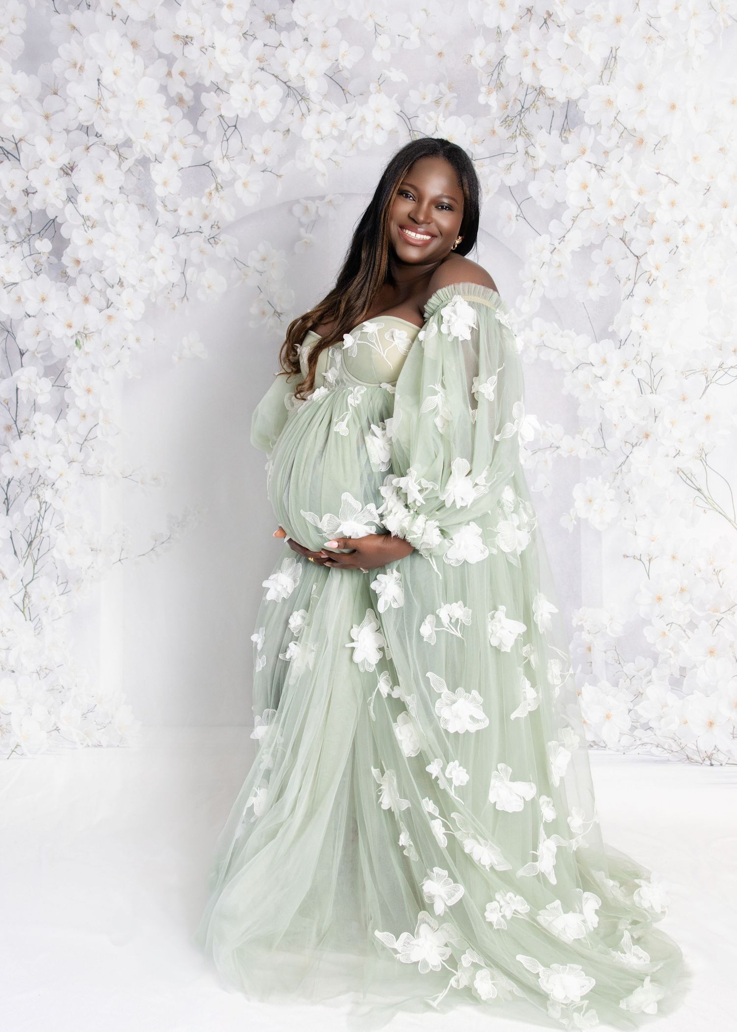 Pregnant woman in sage green floral tulle maternity gown poses gracefully against white floral backdrop.