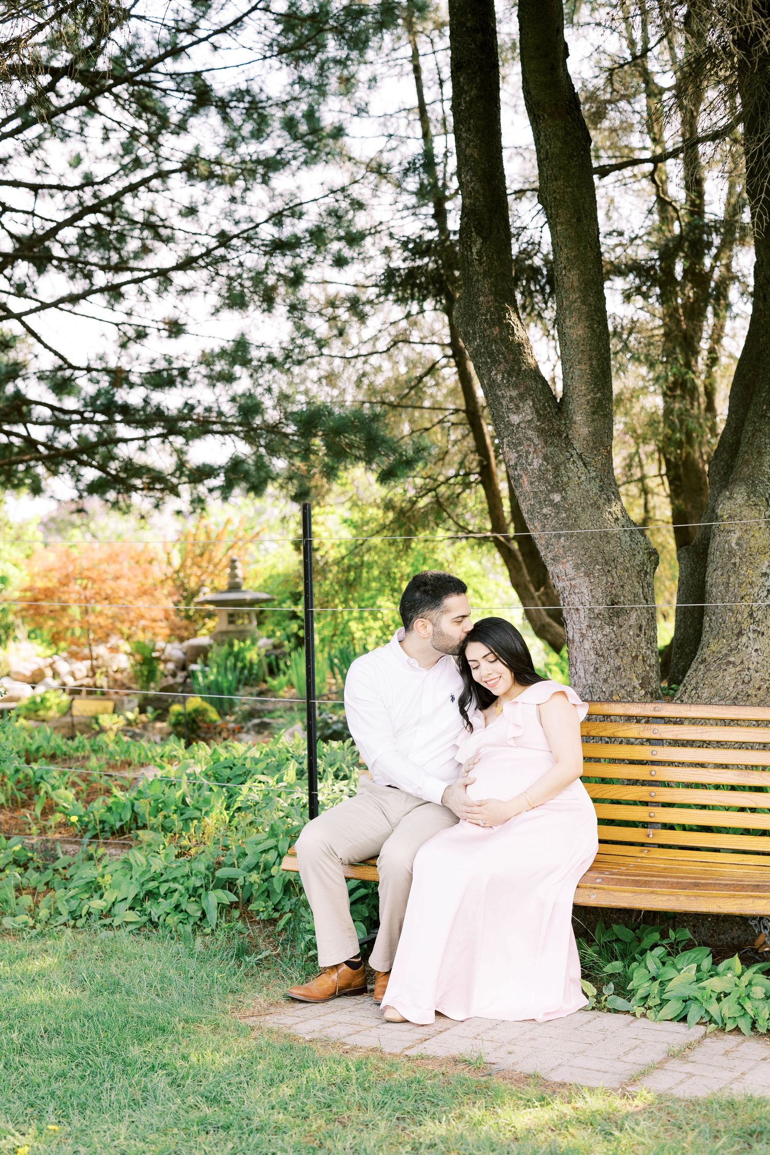 Couple sharing tender moment on wooden bench under large tree, woman in pink maternity gown.
