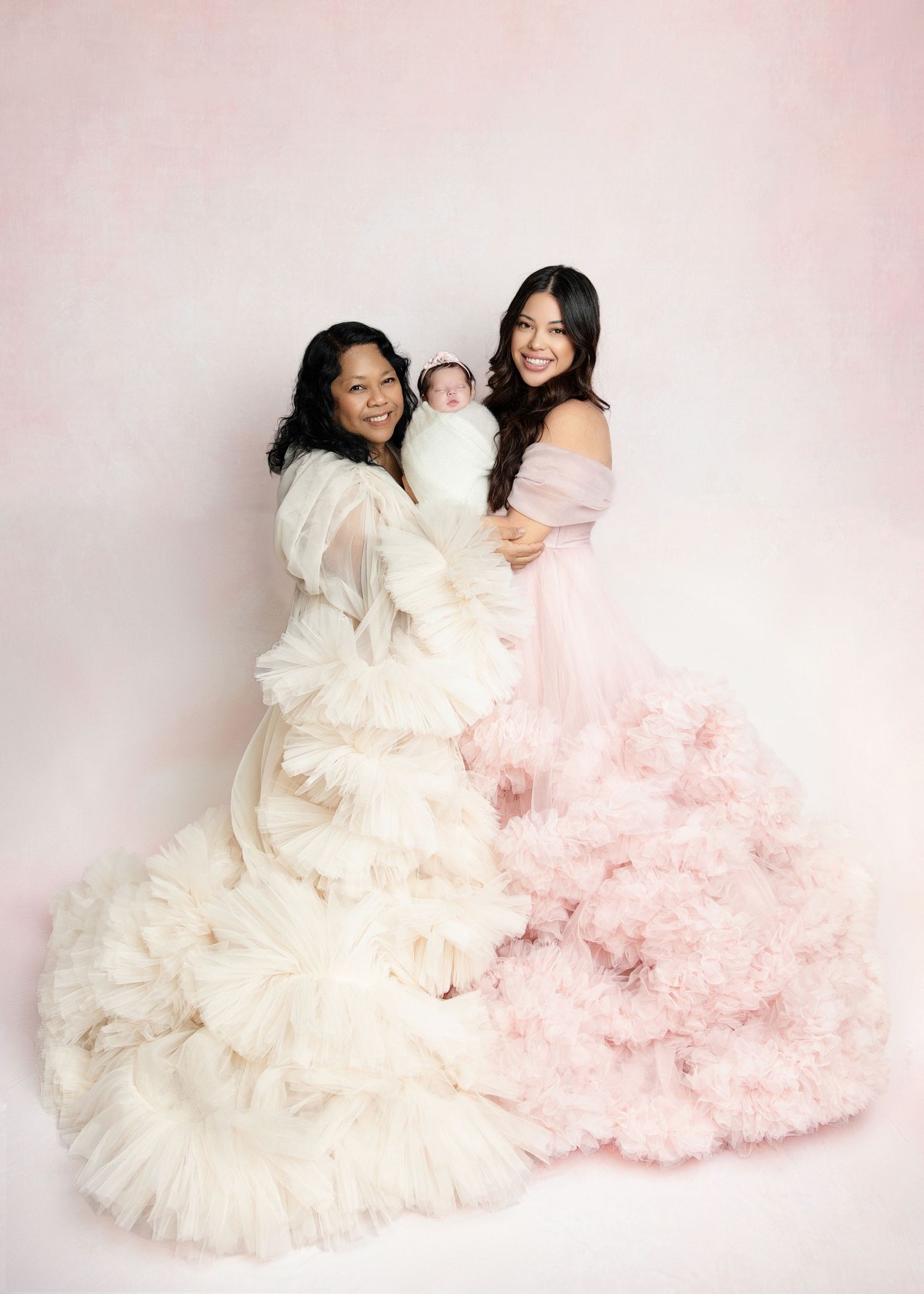 Two women in elegant ruffled gowns, one white and one pink, pose together holding a sleeping newborn baby.