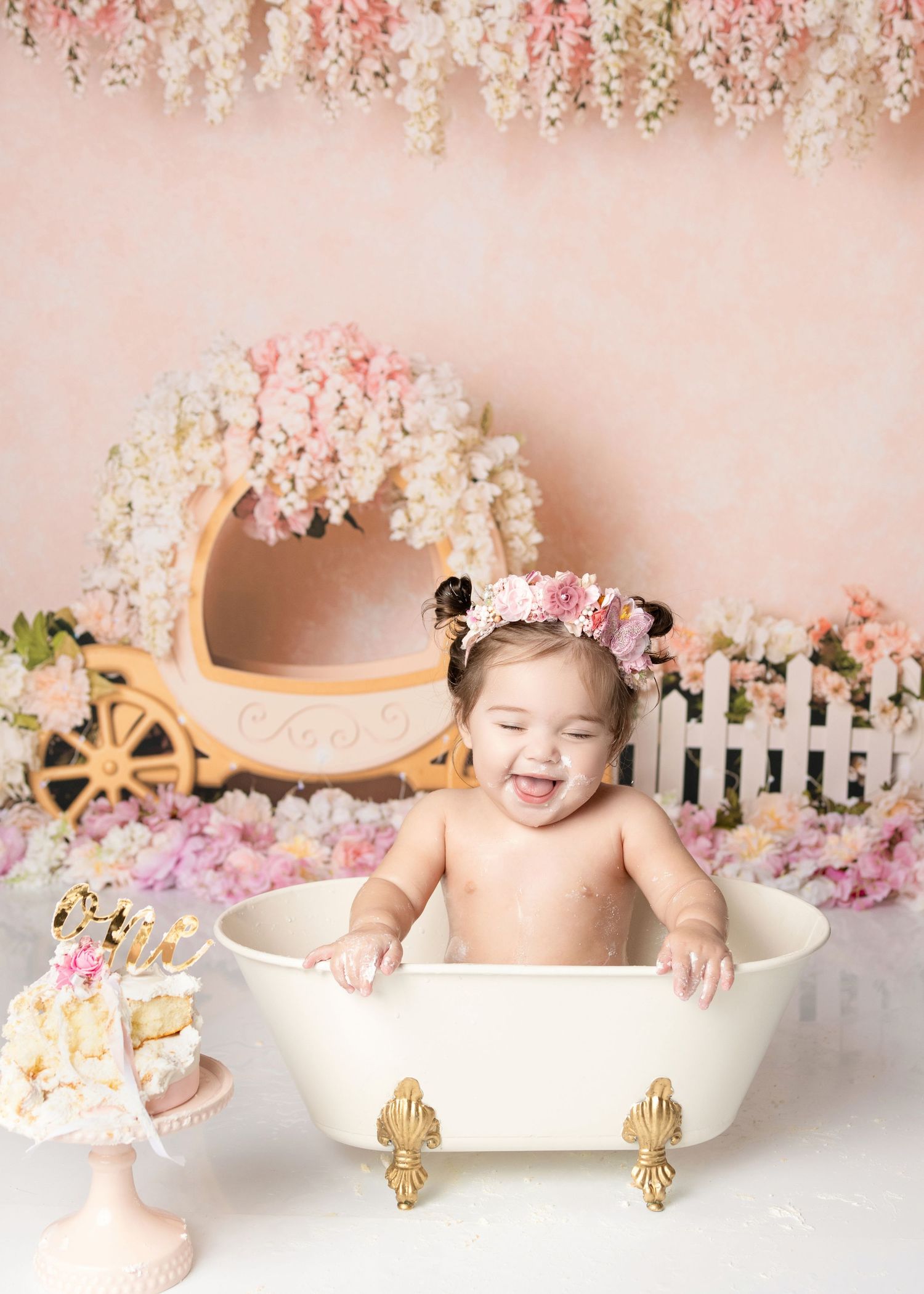 Laughing baby girl wearing floral crown sits in vintage white clawfoot tub at princess first birthday session.