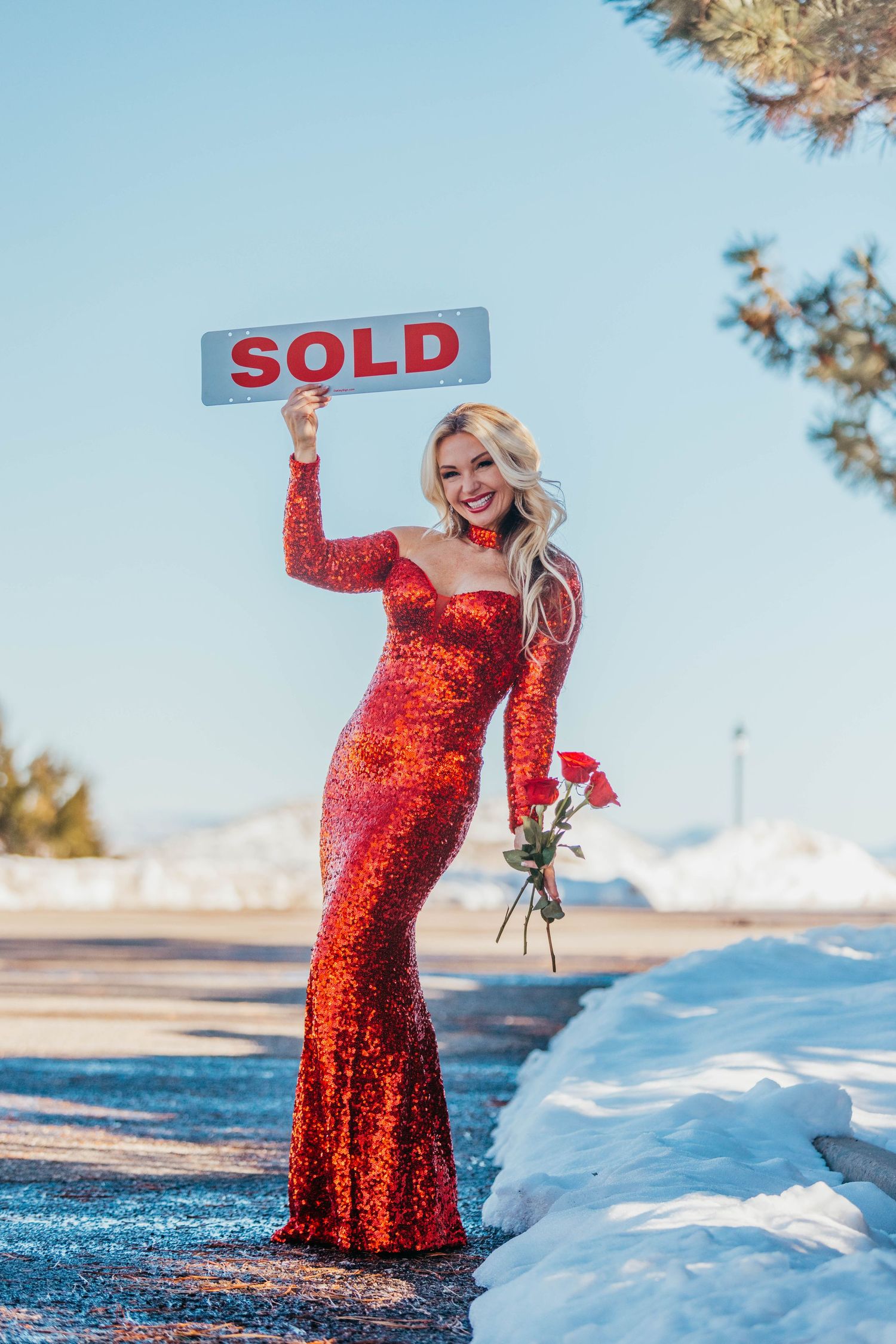 Professional in red sequin dress poses with SOLD sign against snowy landscape.