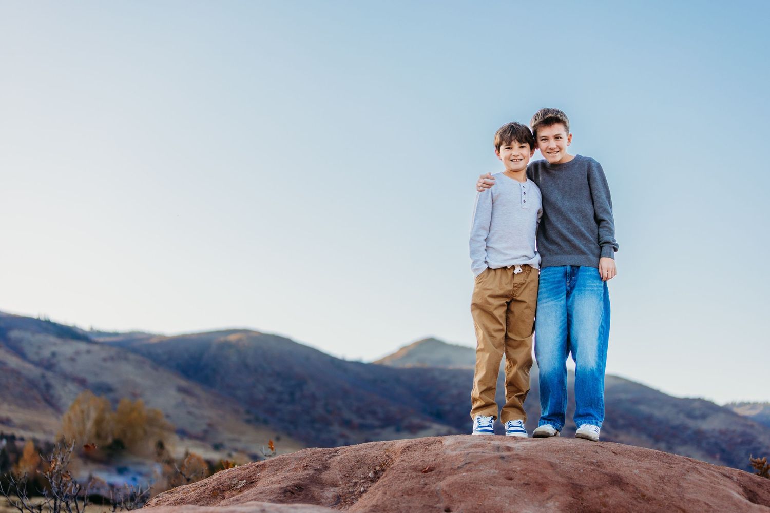 Two boys standing together on a red rock formation with mountain landscape and blue sky backdrop.