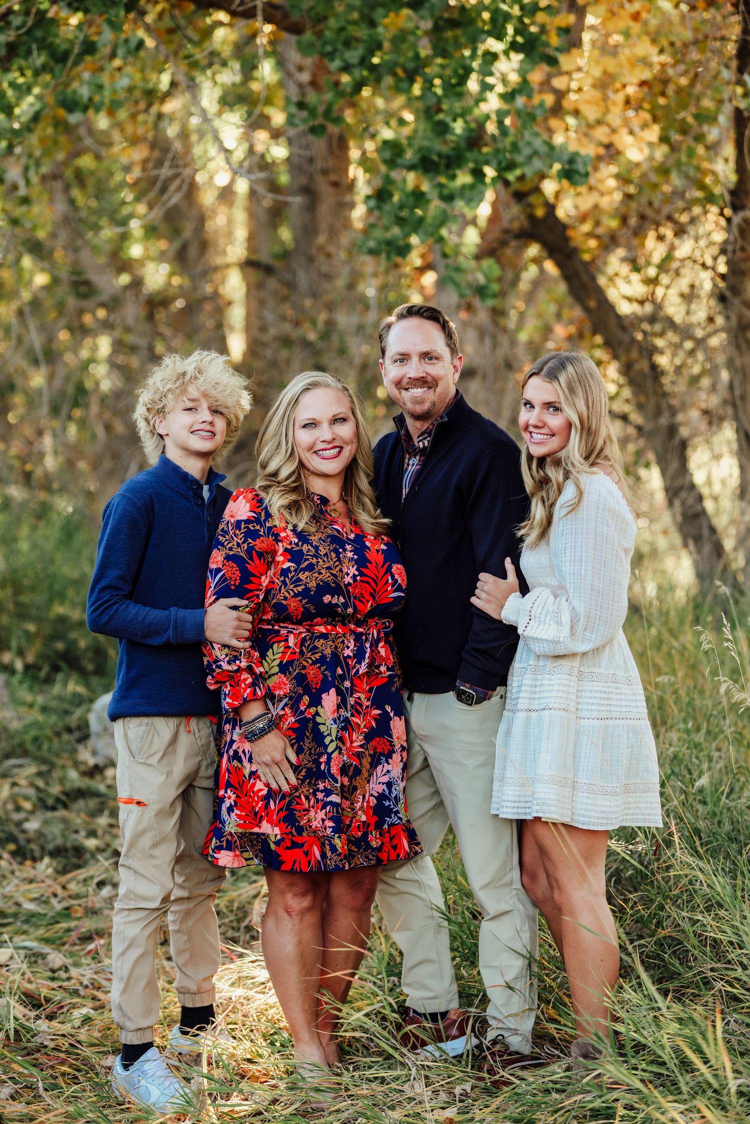 Family of four standing together outdoors in coordinated fall outfits among autumn trees.