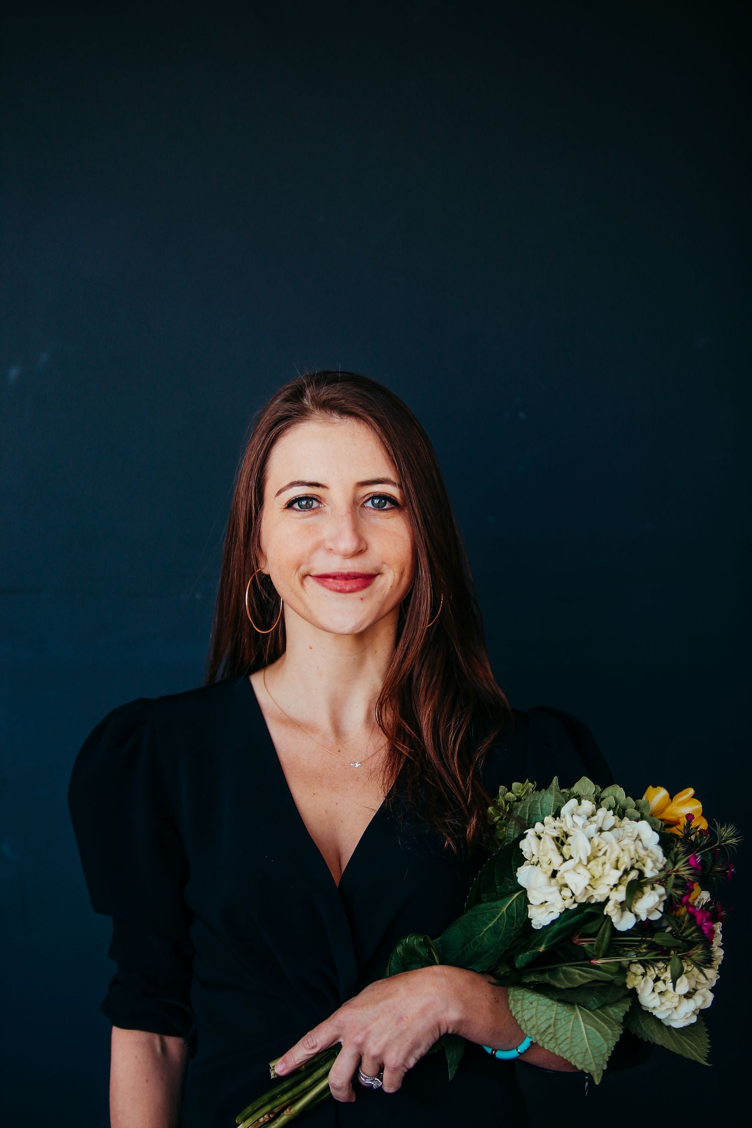 Professional portrait of someone in a black dress holding a bouquet of white hydrangeas against dark background.