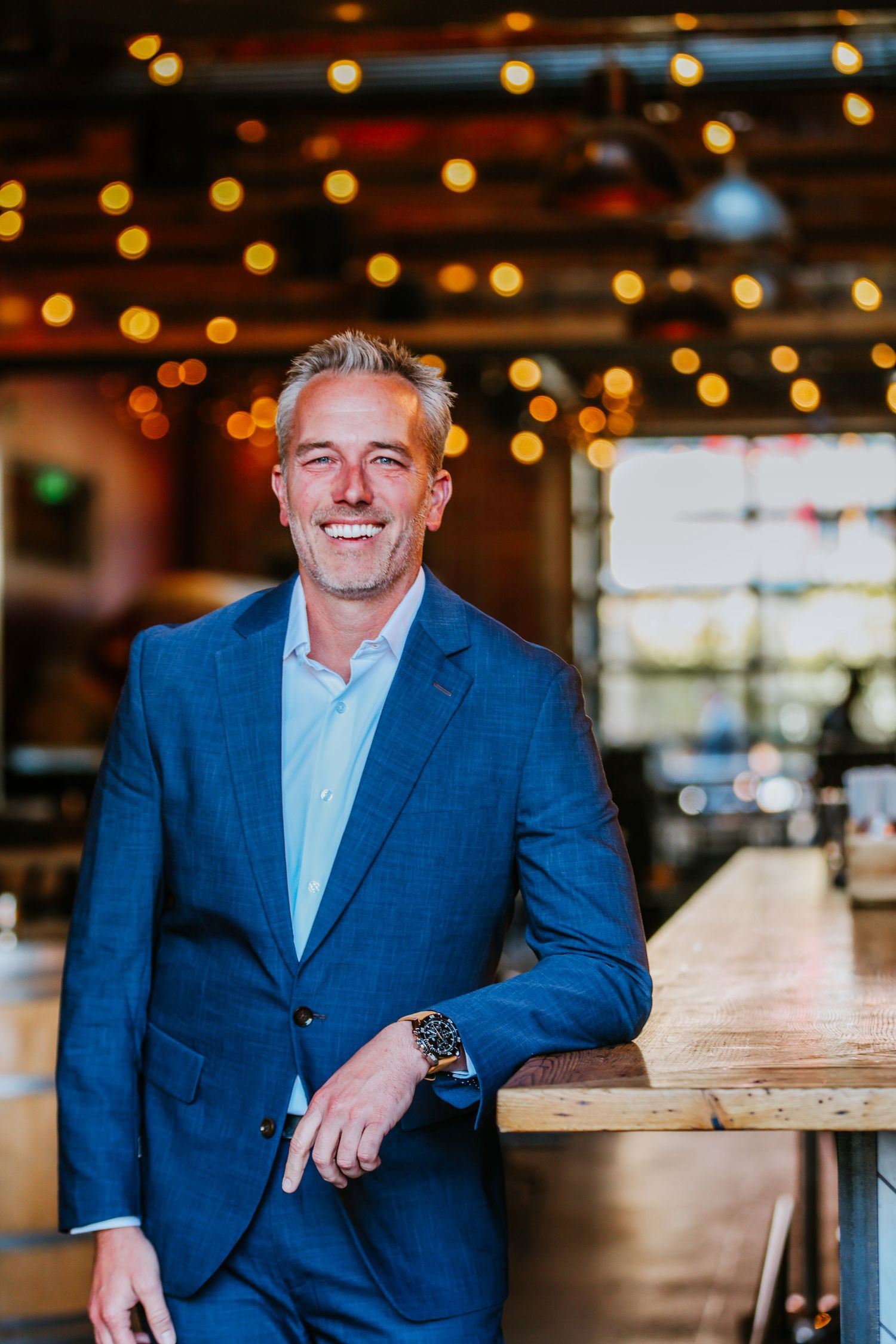 Business professional in navy suit standing by wooden table with warm lighting and bokeh background.