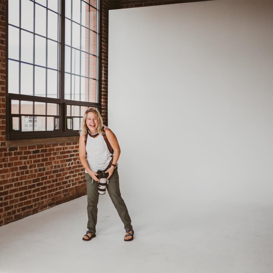 Candid studio portrait with exposed brick and windows showing photographer holding camera.
