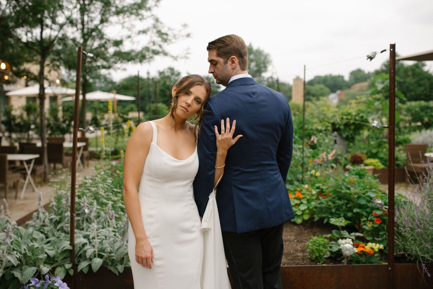 Bride and groom pose in a lush garden wedding venue, she in white gown, he in navy suit.