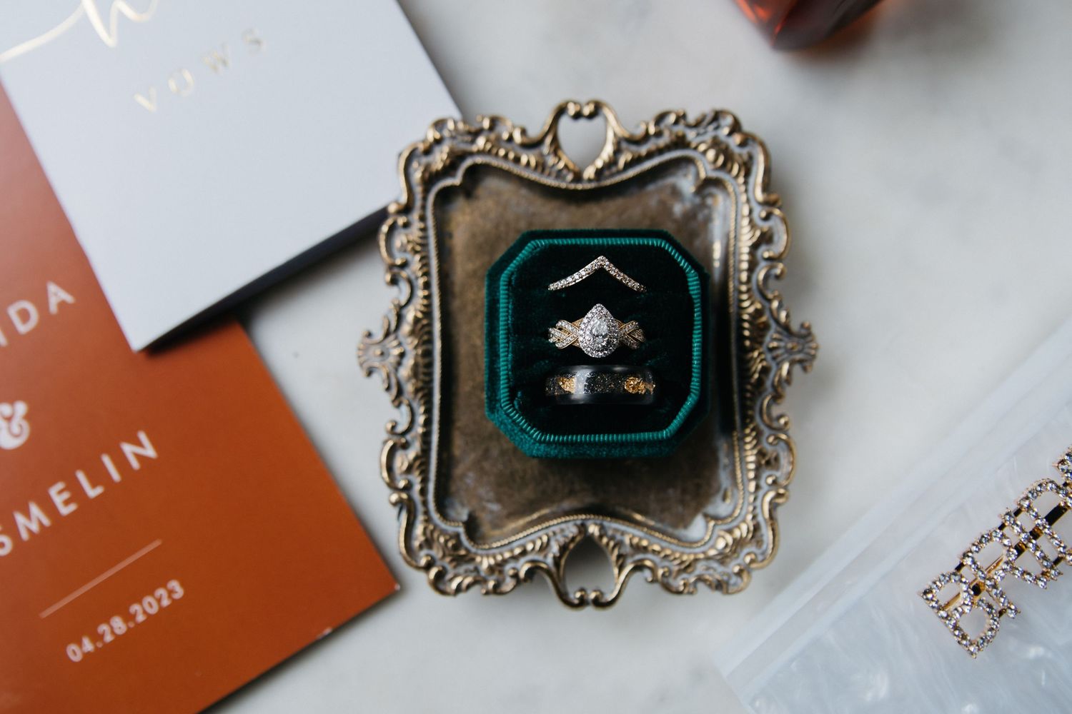 Elegant wedding rings displayed in a green velvet box on an ornate silver tray beside wedding stationery dated 04.28.2023.