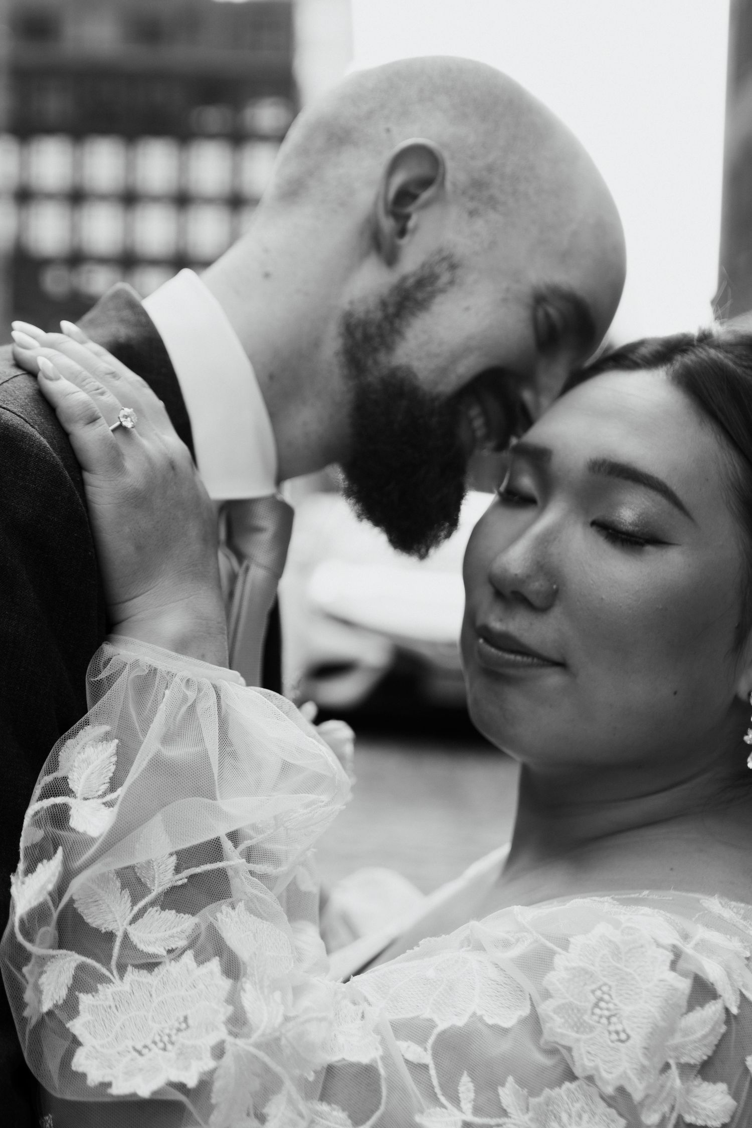 Black and white wedding photo of a bald bearded groom leaning forehead to bride in lace dress.