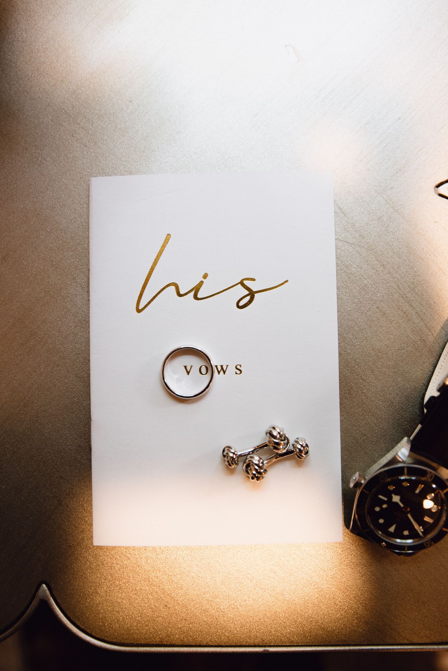 Gold wedding ring and cufflinks on a white his vows card beside a watch on a golden surface.