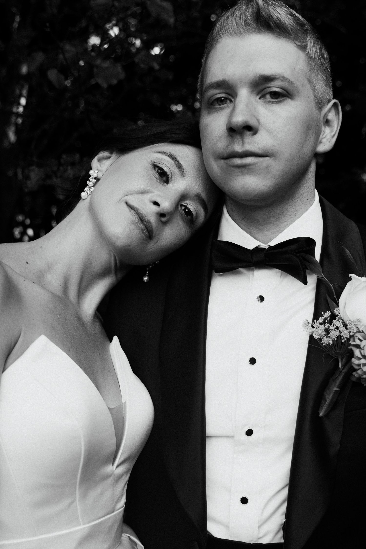 Black and white wedding portrait of bride in white gown leaning on groom in tuxedo with boutonniere.