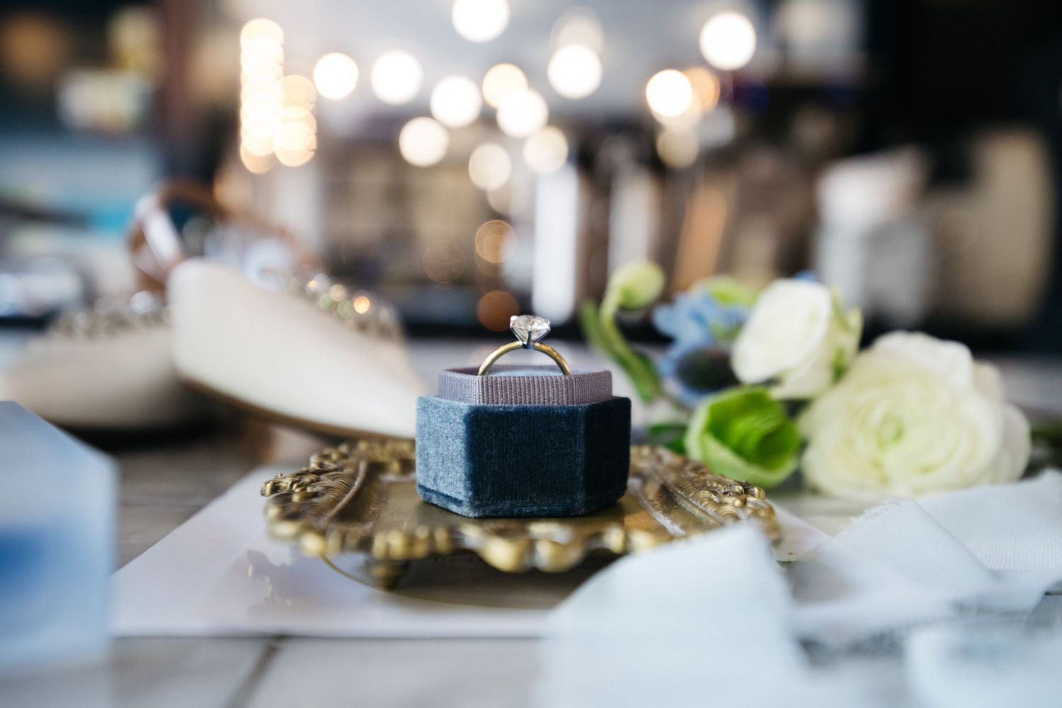 Diamond engagement ring in a velvet box on a gold tray with white roses and bokeh lights in background.