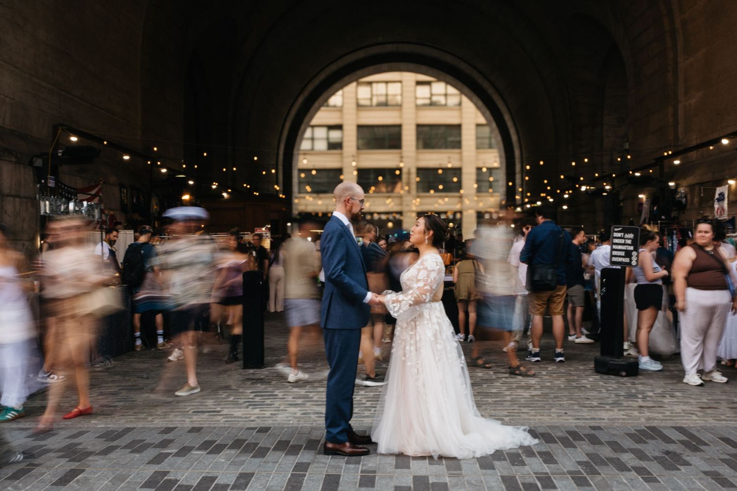 Bride and groom share a romantic moment under a grand arch with string lights and blurred wedding guests in the background.