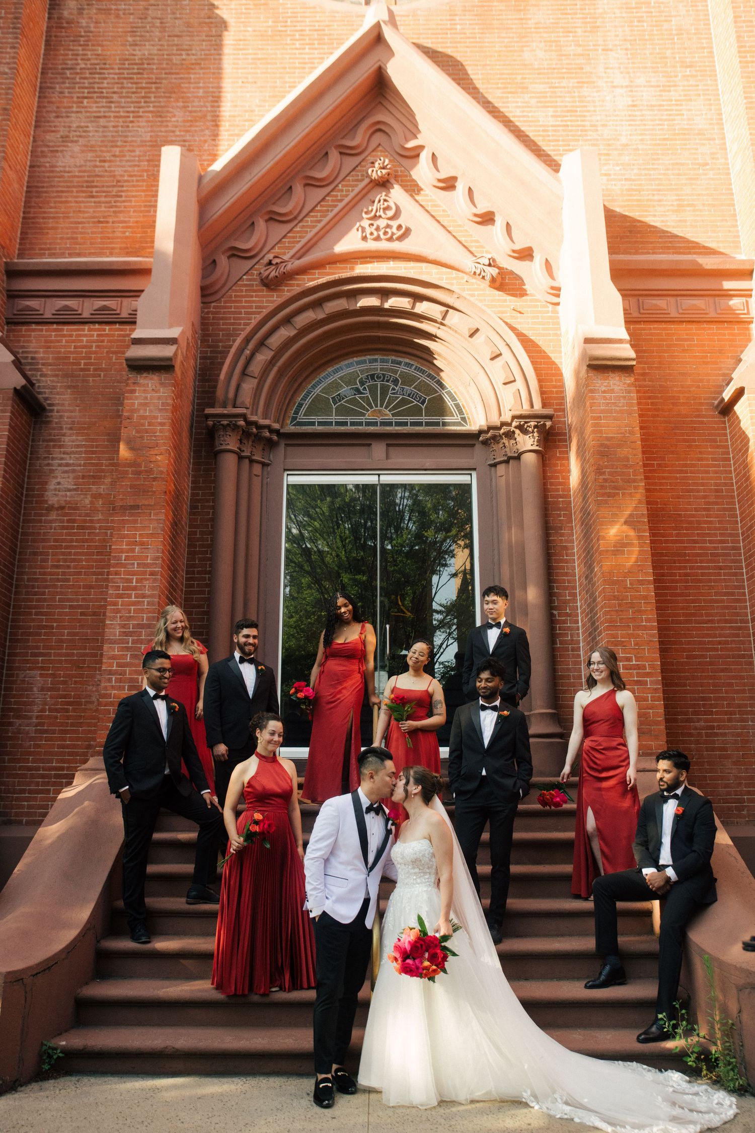 Wedding party in red and black formal wear poses on brick church steps, bride and groom share a kiss in front.