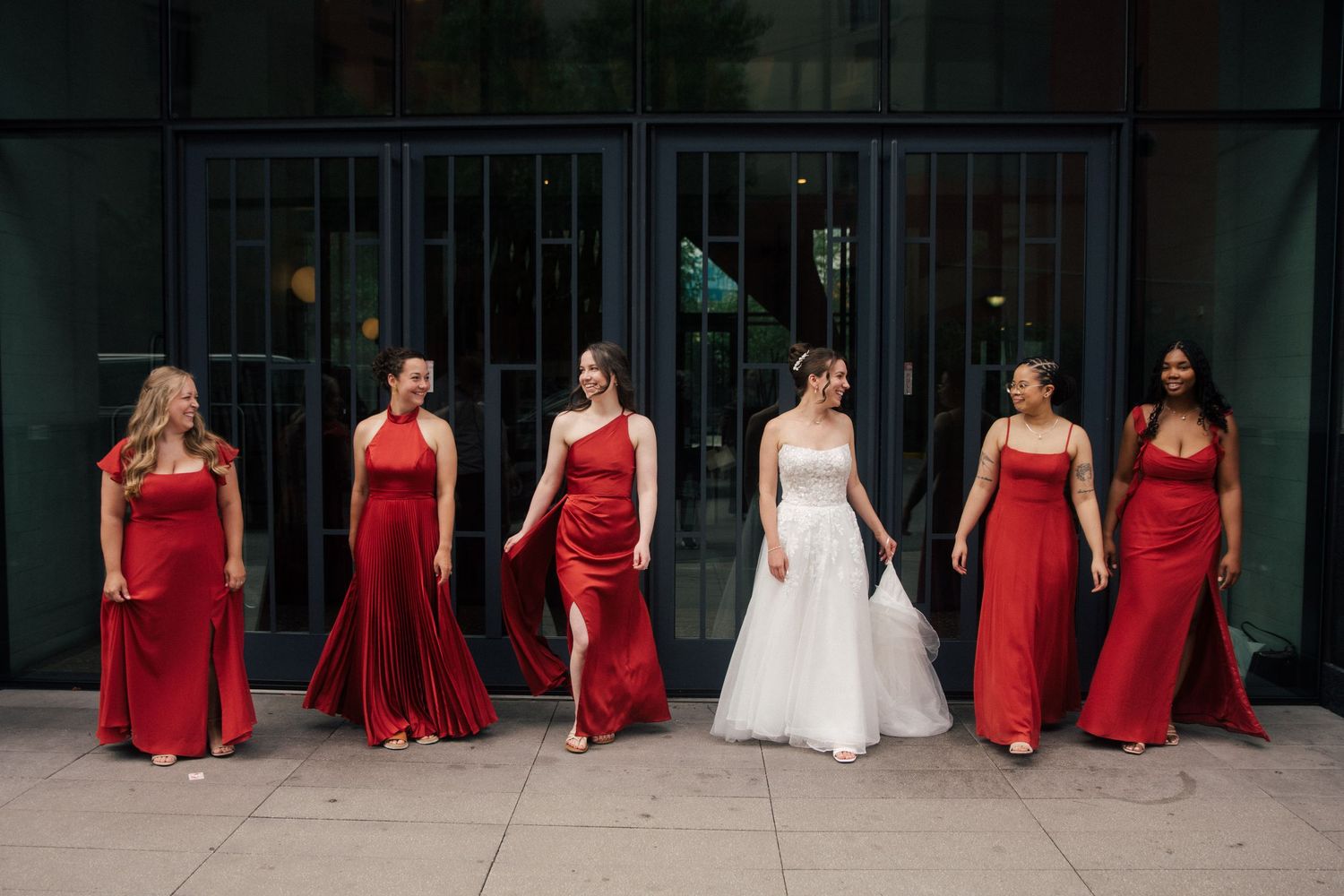 Bride in white gown walks with six bridesmaids in red dresses outside a modern building.