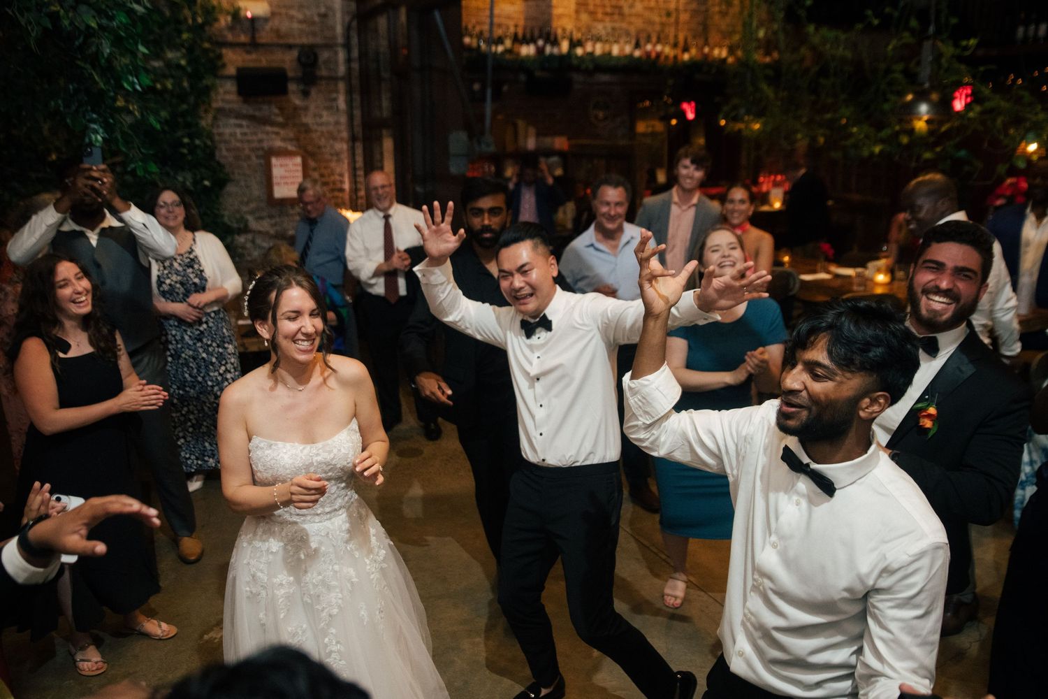Joyful wedding reception with bride in white gown, groom in tuxedo, and guests dancing and celebrating indoors.
