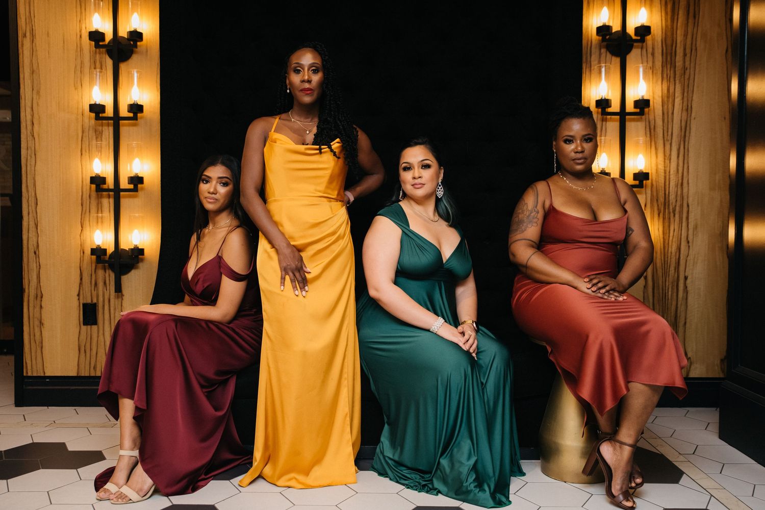Four women in elegant satin gowns in jewel tones pose gracefully in a luxurious venue setting.