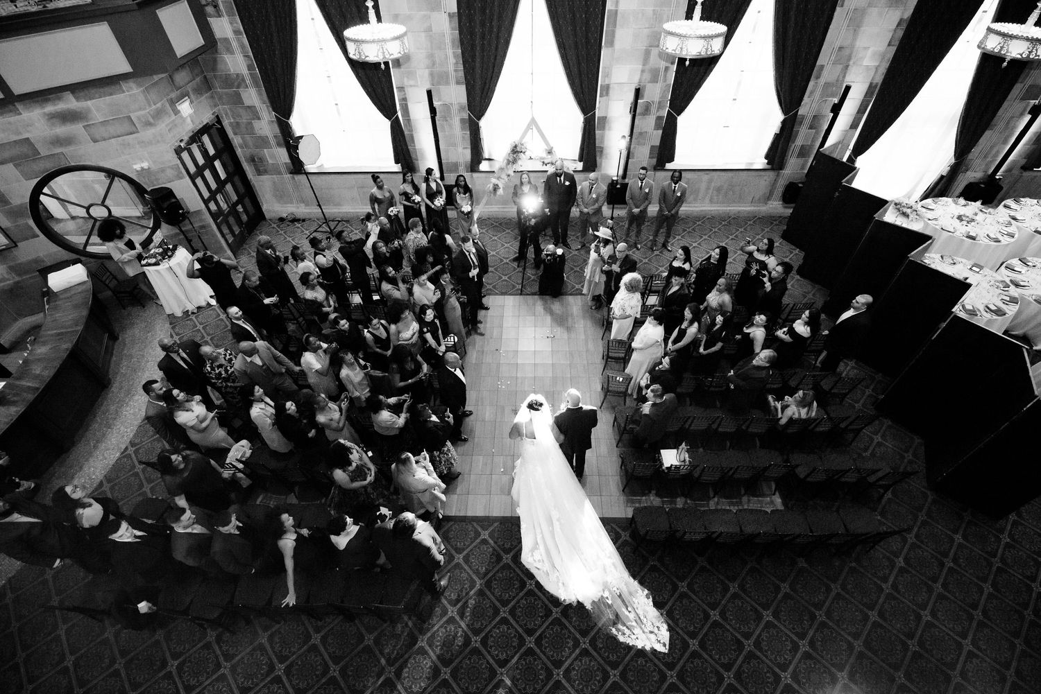 Black and white aerial view of a wedding ceremony in an elegant venue with guests seated in pews and bride walking the aisle.