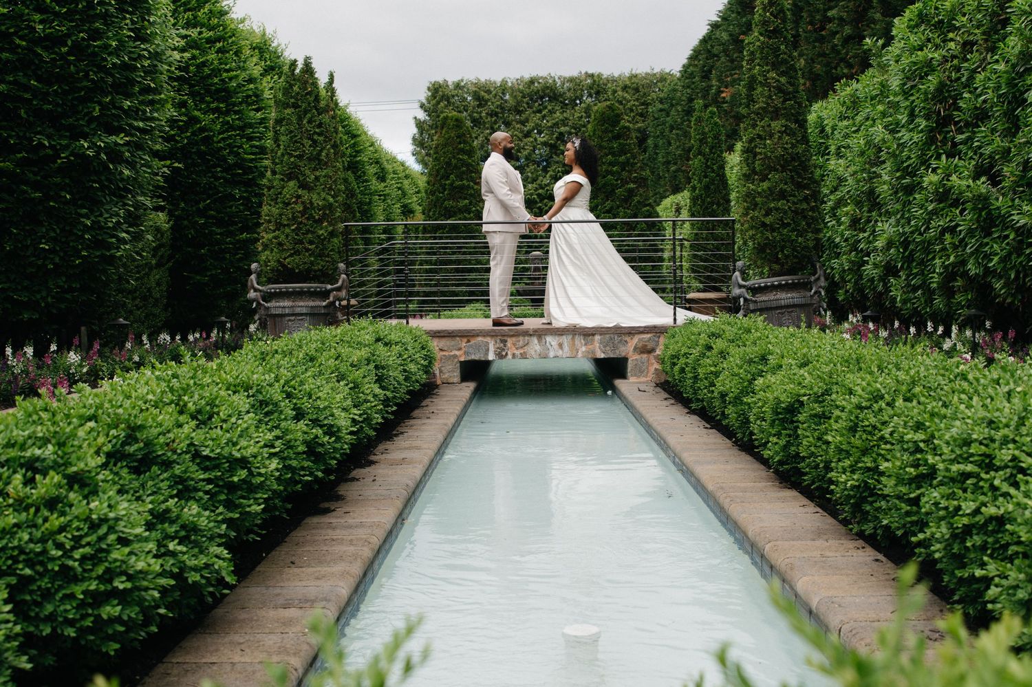 Bride and groom stand on a bridge over a reflecting pool in a lush formal garden with manicured hedges.