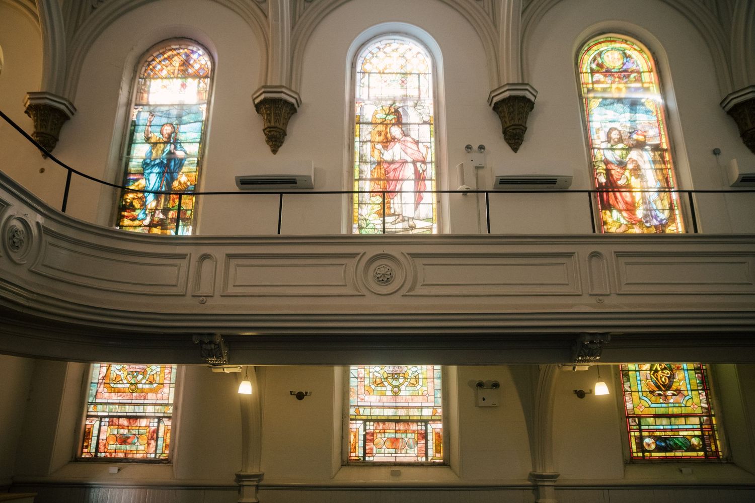 Church interior with ornate stained glass windows depicting religious figures, white walls, and a curved balcony.