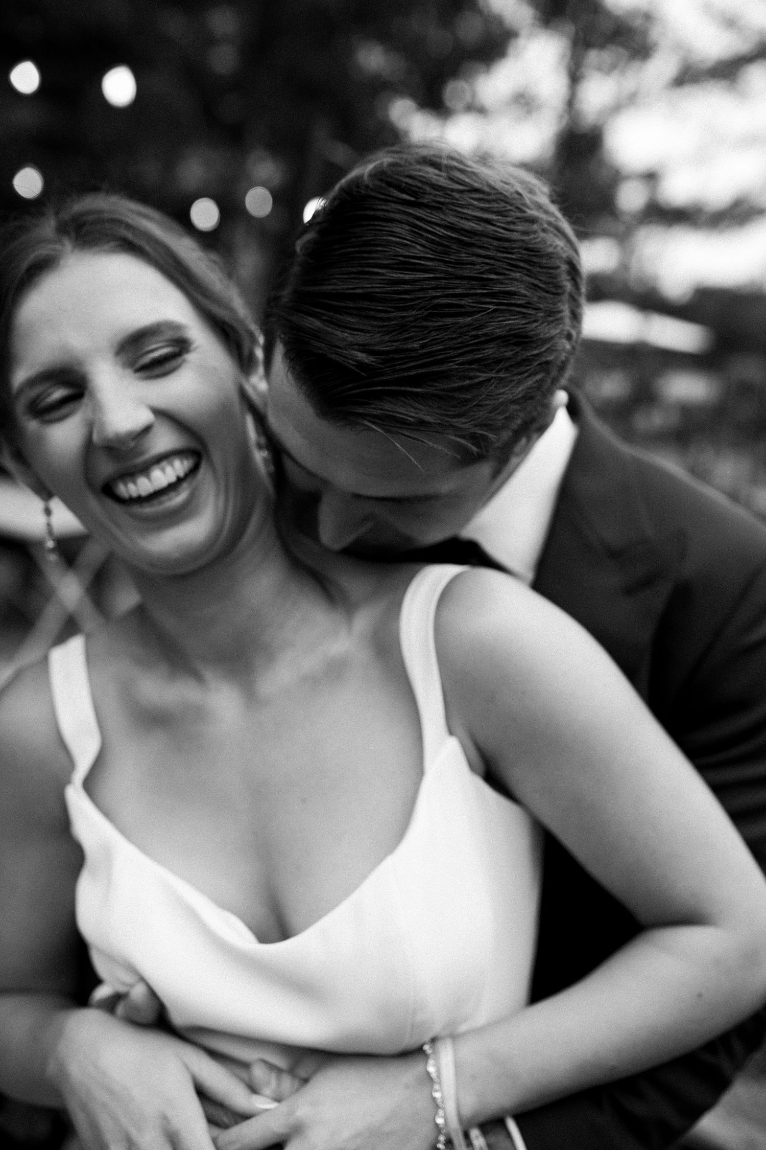 Black and white photo of a laughing bride in a white dress being embraced by a groom in a dark suit.
