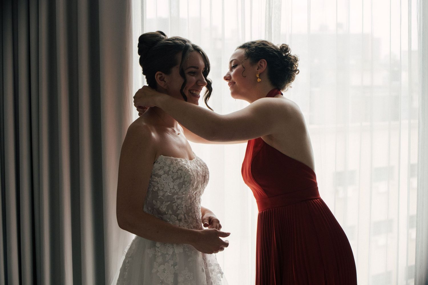 Bride in white lace gown smiles as bridesmaid in red dress fastens her necklace near a bright window.