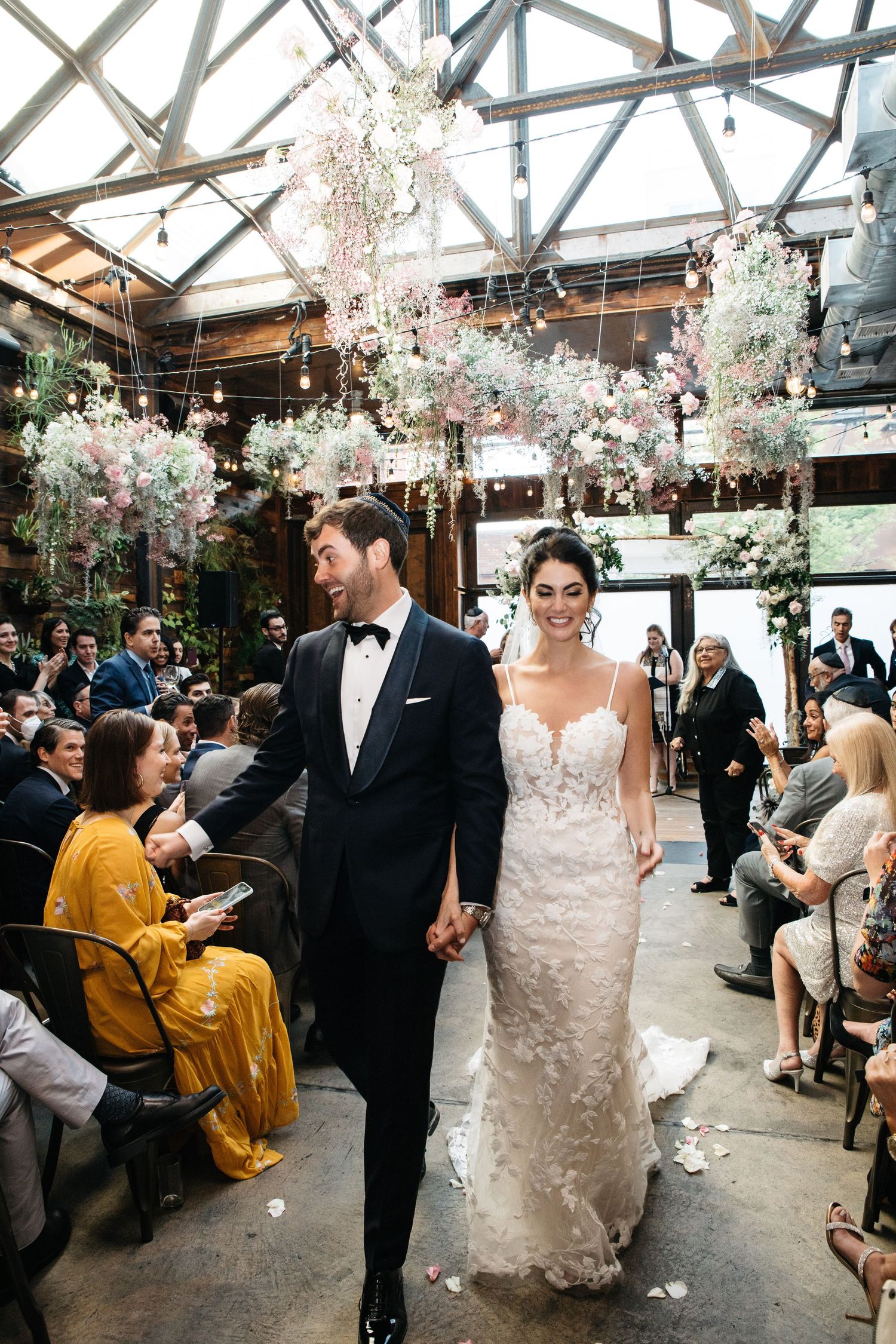 Bride and groom walk down the aisle hand in hand beneath floral installations at an elegant indoor wedding ceremony.