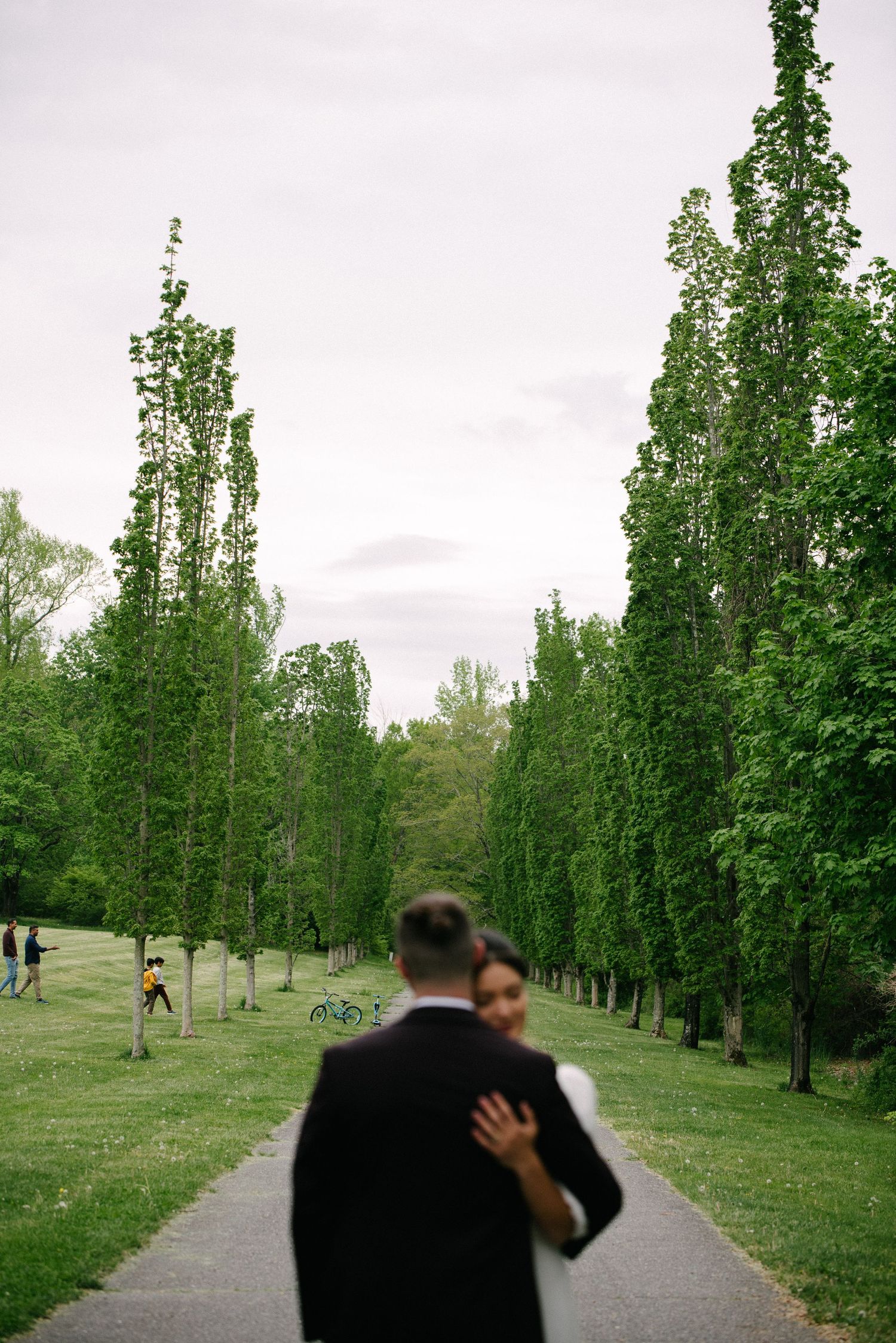 Couple embracing on a tree-lined park path, groom in black suit, lush green surroundings on a cloudy day.