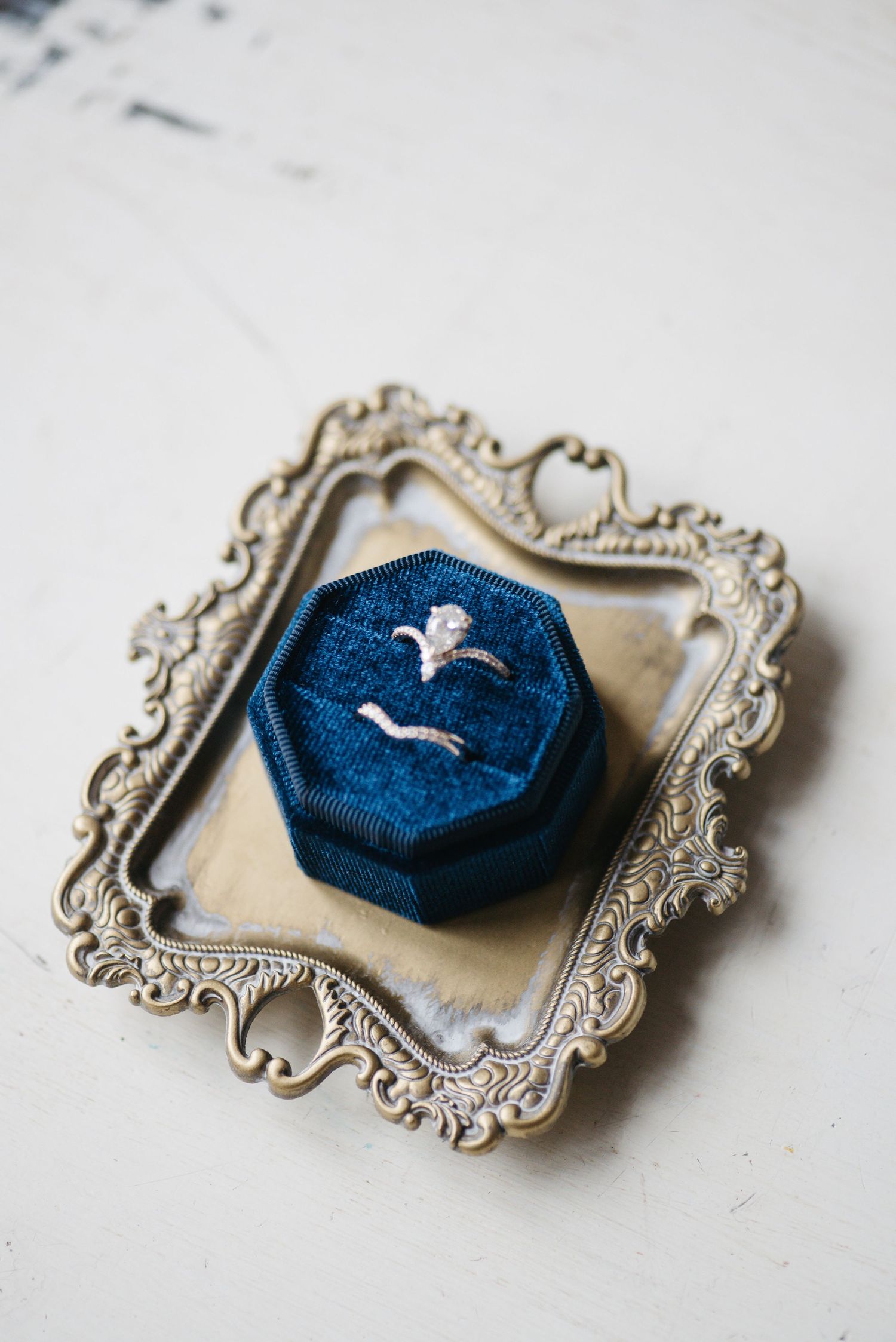 Blue velvet octagonal ring box with diamond engagement rings on ornate gold vintage tray against white background.