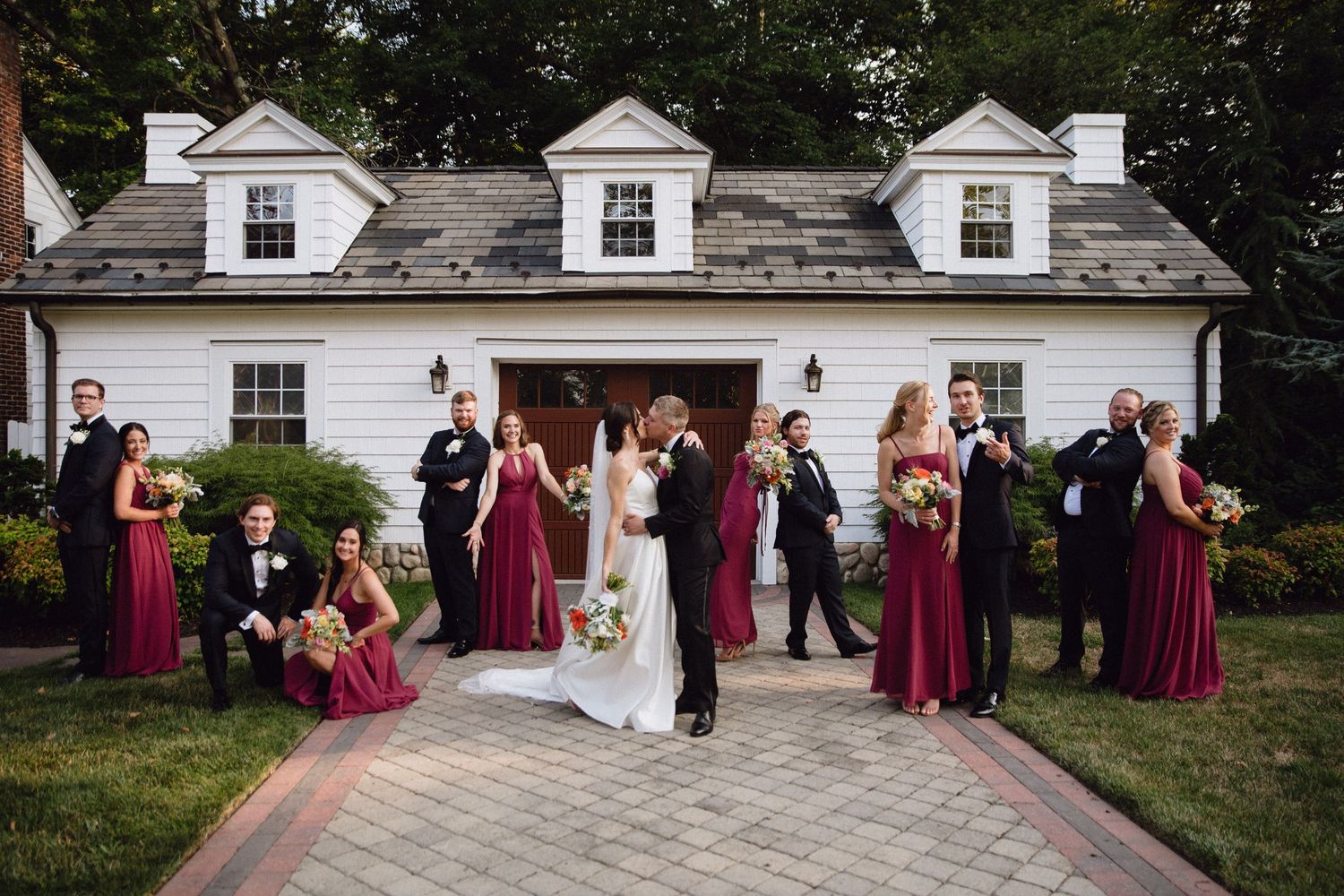 Wedding party in burgundy and black attire poses outside a white colonial venue with the bride and groom kissing.